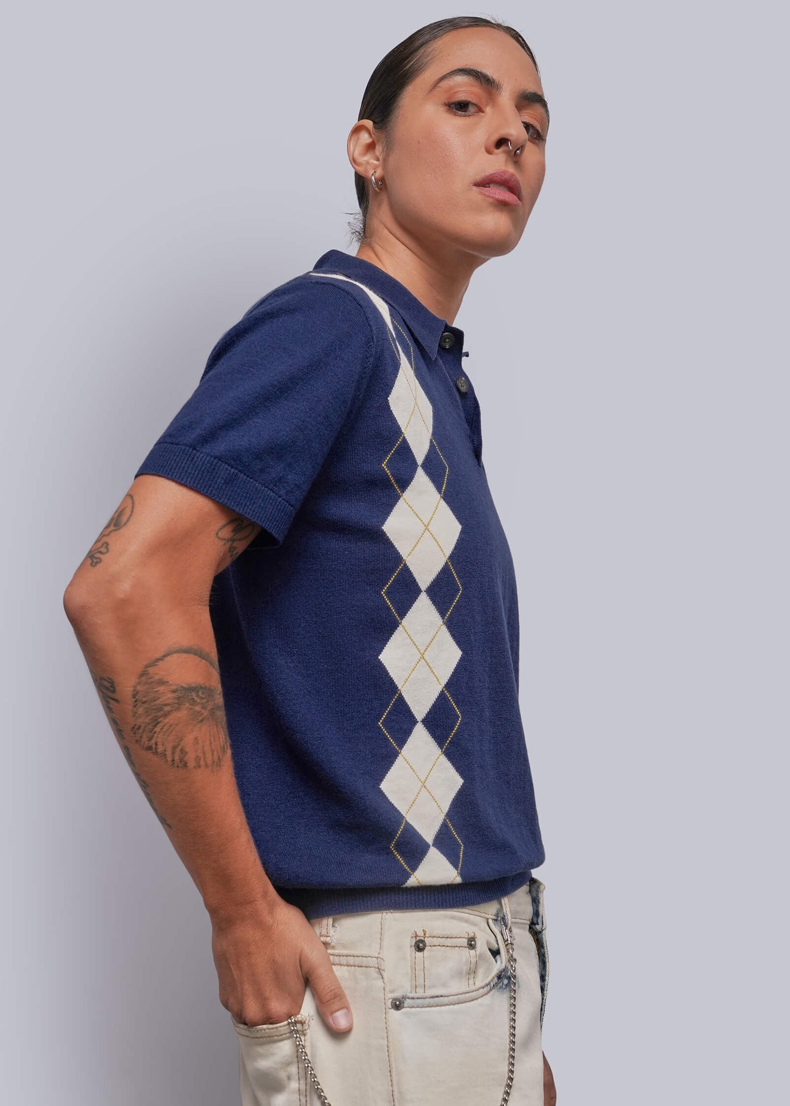 S=A model wearing a navy blue short-sleeve knit polo shirt with a cream argyle pattern on the front, paired with beige pants, posed against a solid light gray background.