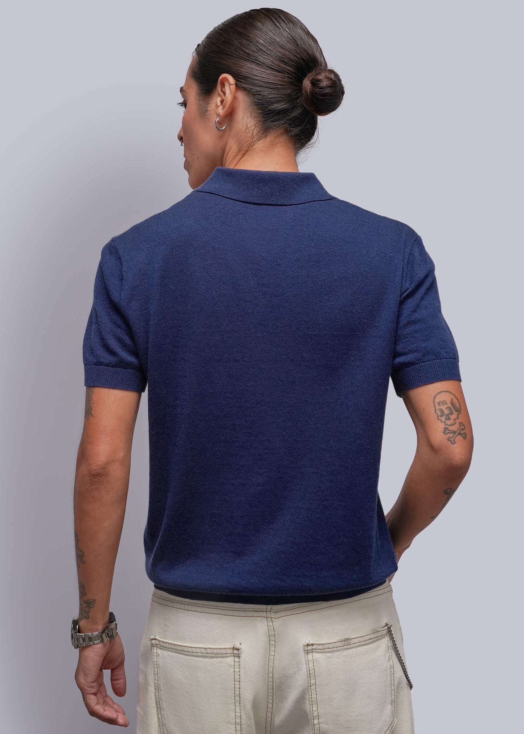 S=A person shown from the back wearing a navy blue short-sleeve polo shirt and light beige trousers, standing against a solid gray studio background.