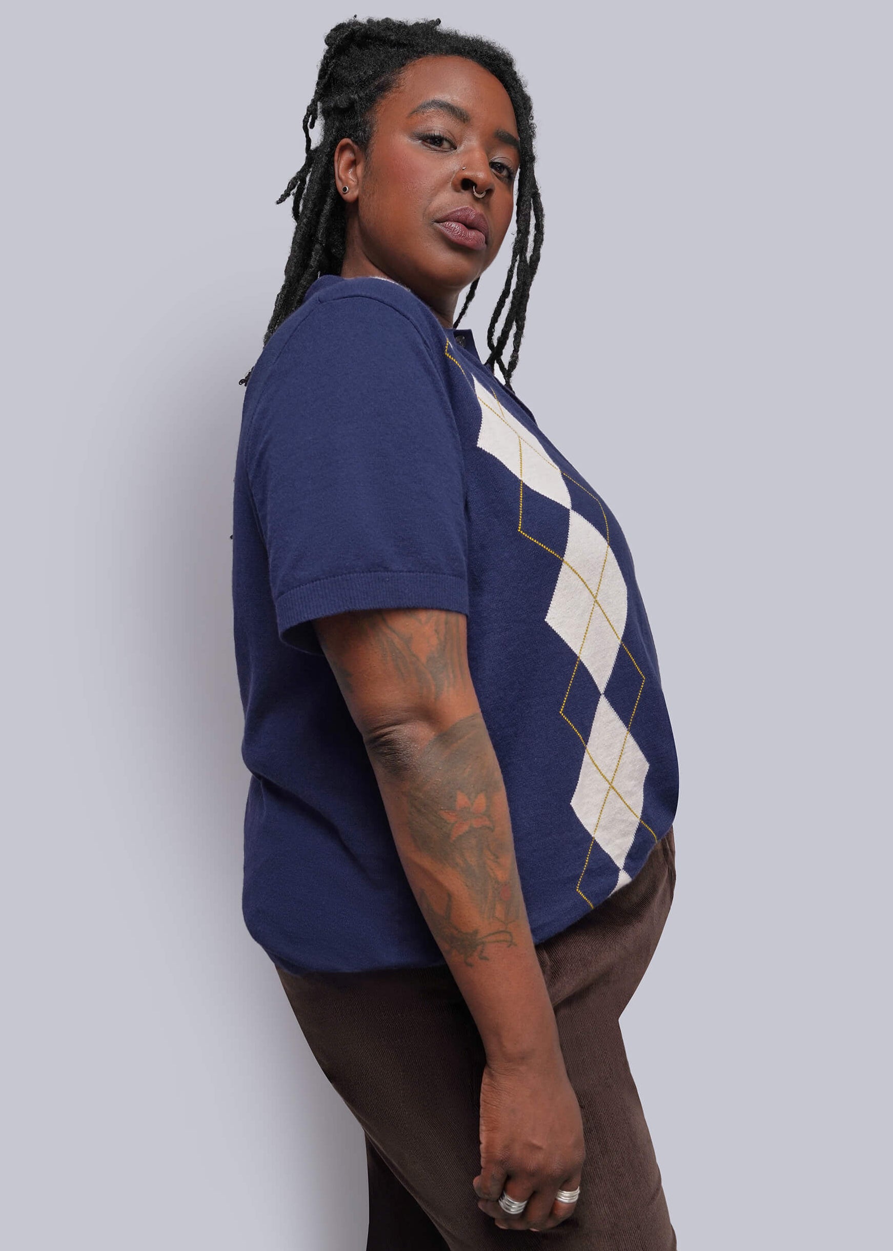 1X=A woman wearing a navy blue short-sleeve t-shirt with a cream and gold argyle pattern on the front, paired with brown pants, standing against a solid light gray background.