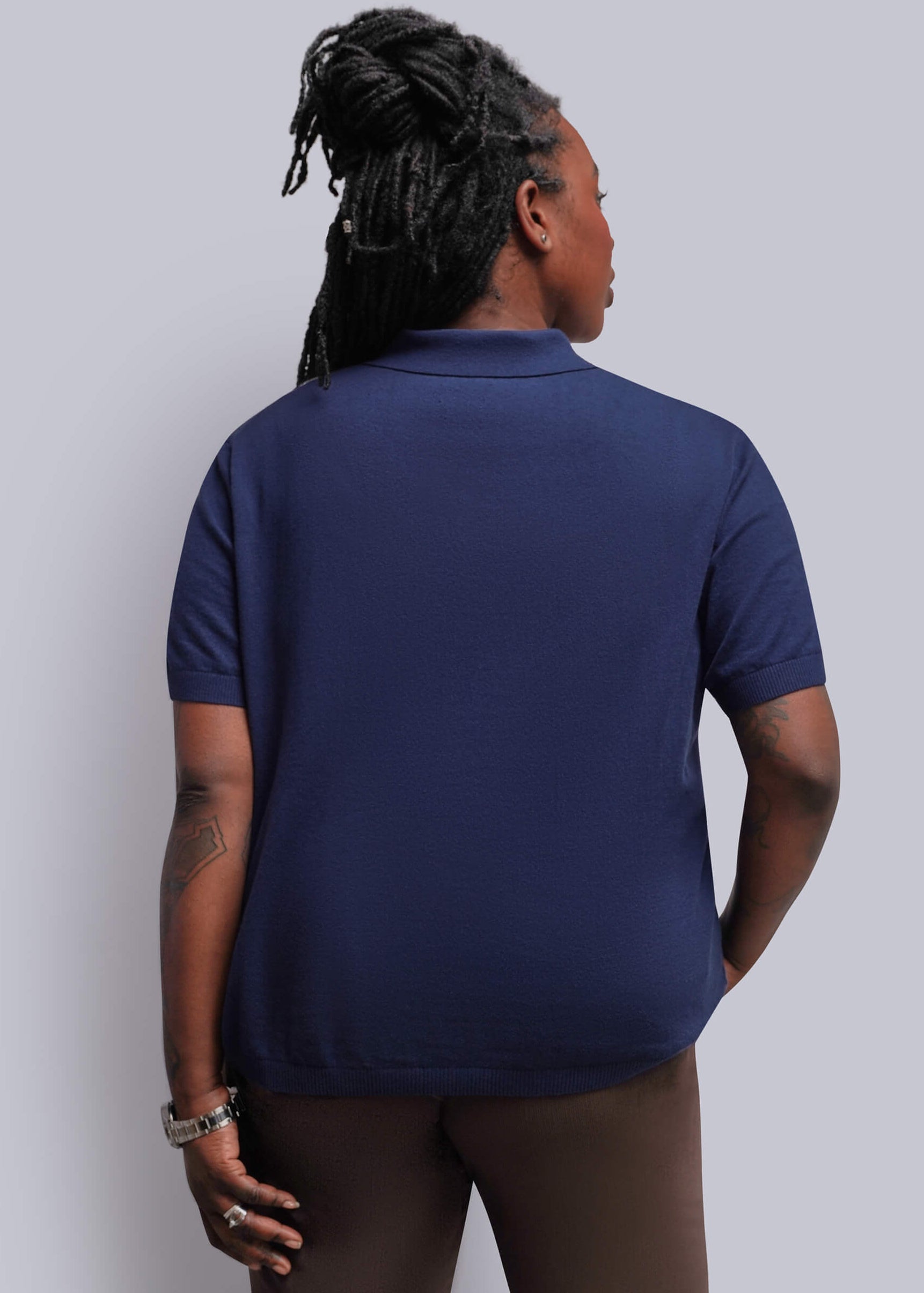 1X=A woman shown from the back wearing a navy blue short-sleeve polo shirt and dark brown pants, standing against a solid light gray studio background.