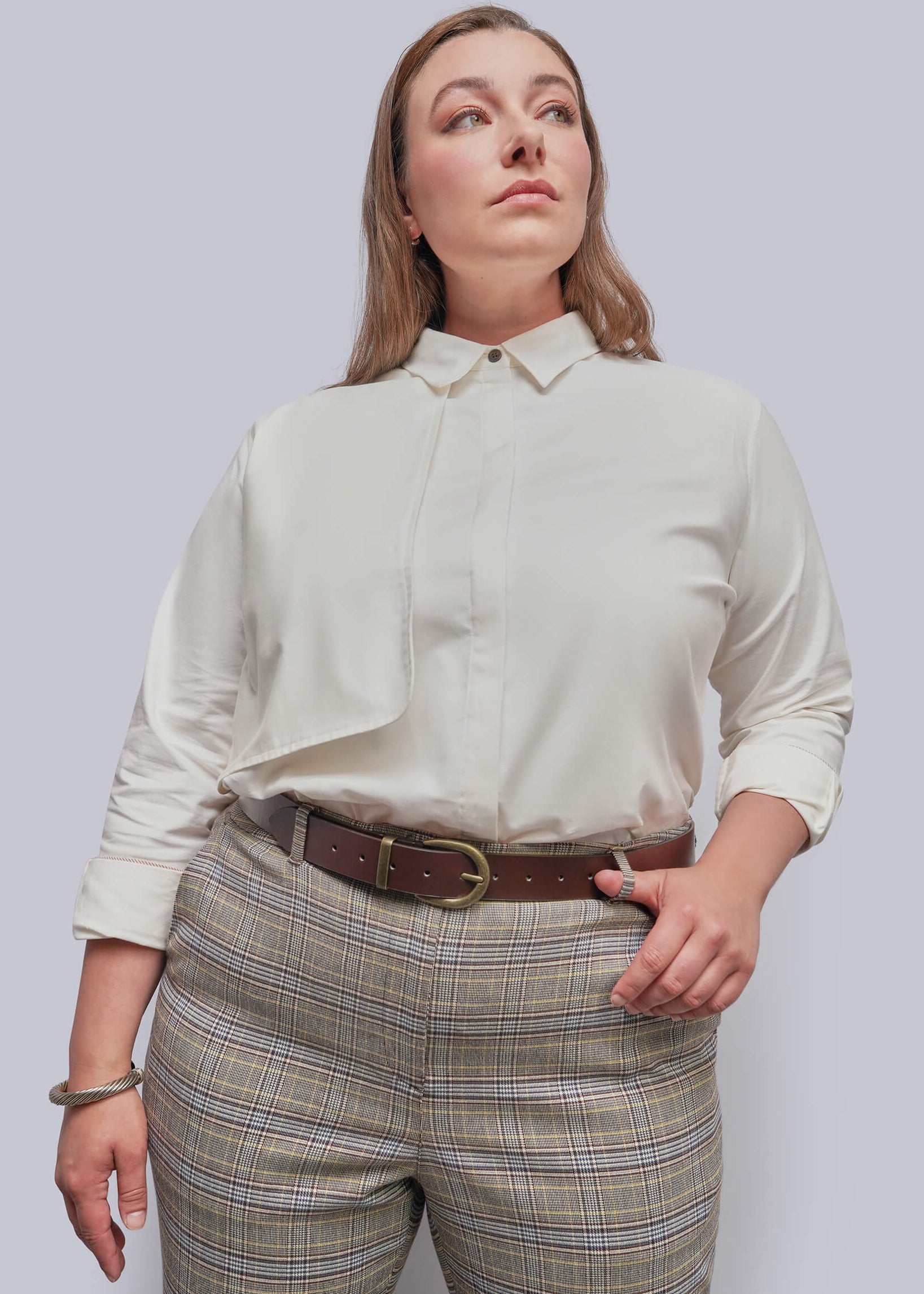 A woman wearing a cream-colored long-sleeve button-down shirt tucked into beige plaid trousers with a brown belt, photographed against a solid light gray background.