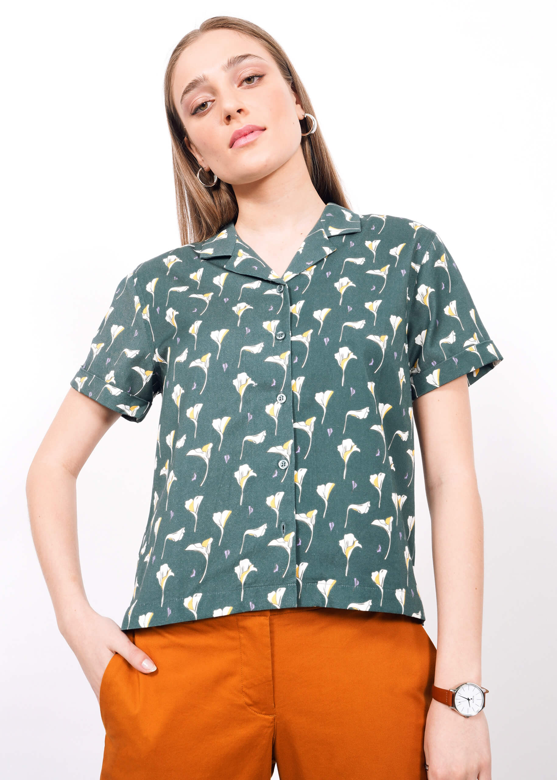 S= A woman with straight, light brown hair wears The Everyday Camp Button Up—a green short-sleeved shirt with a white floral pattern—paired with orange pants, hoop earrings, and a wristwatch, standing against a plain white background.