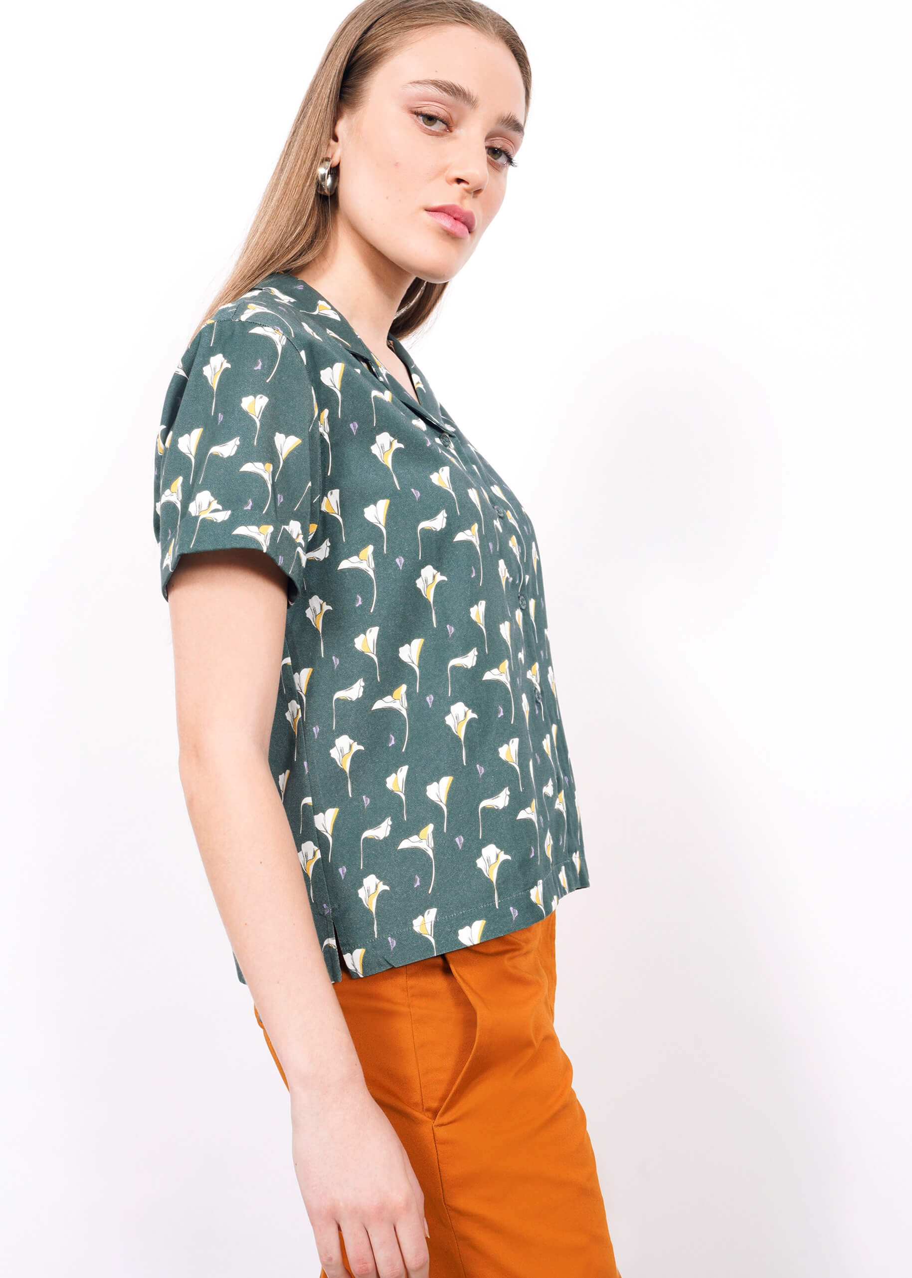 S=A woman wears The Everyday Camp Button Up in green floral print with orange pants, standing against a plain white background and looking at the camera with a neutral expression.