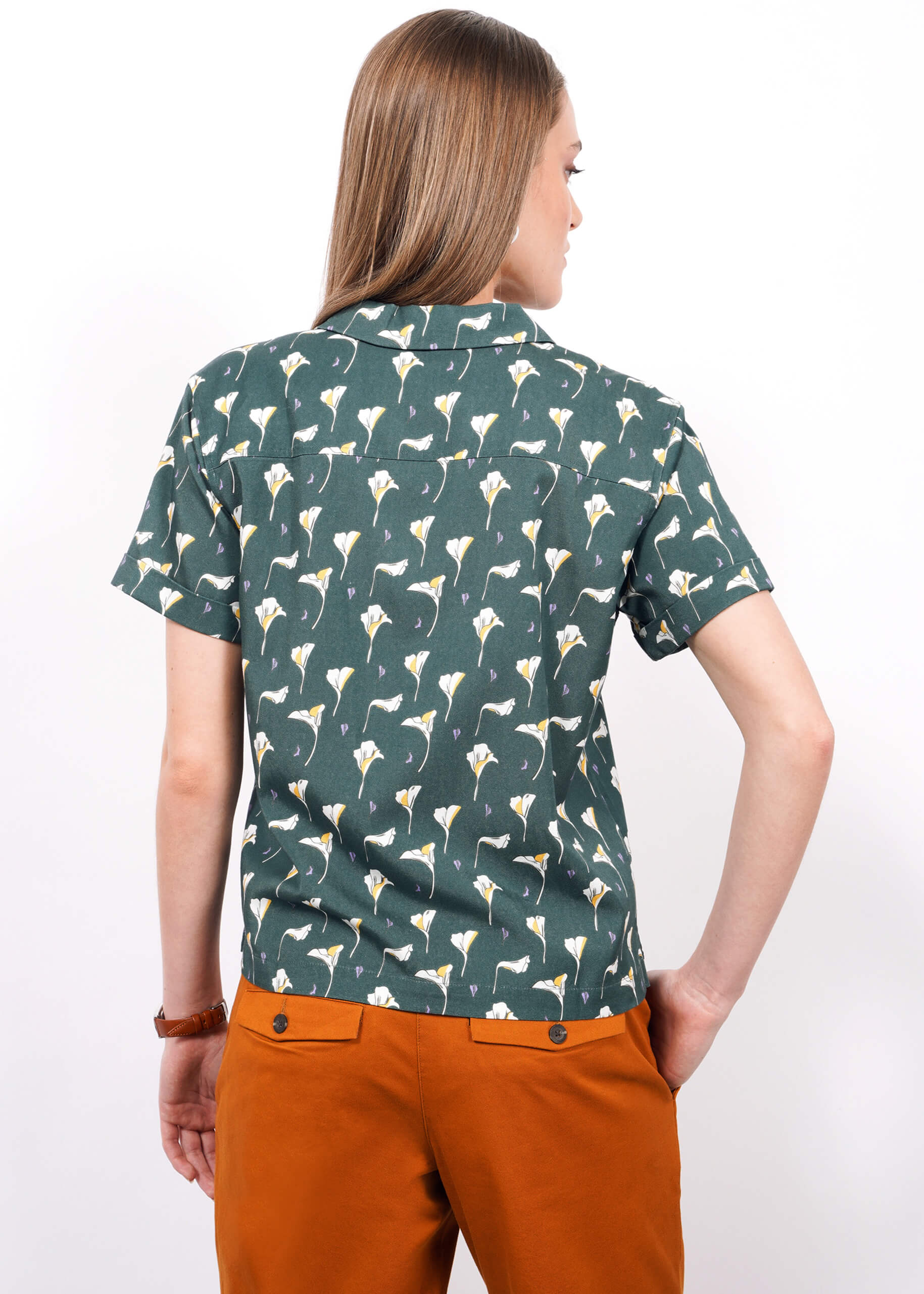 S=A person with straight, light brown hair wears The Everyday Camp Button Up in green Tencel with a white floral pattern and orange pants, shown from the back against a plain white background.