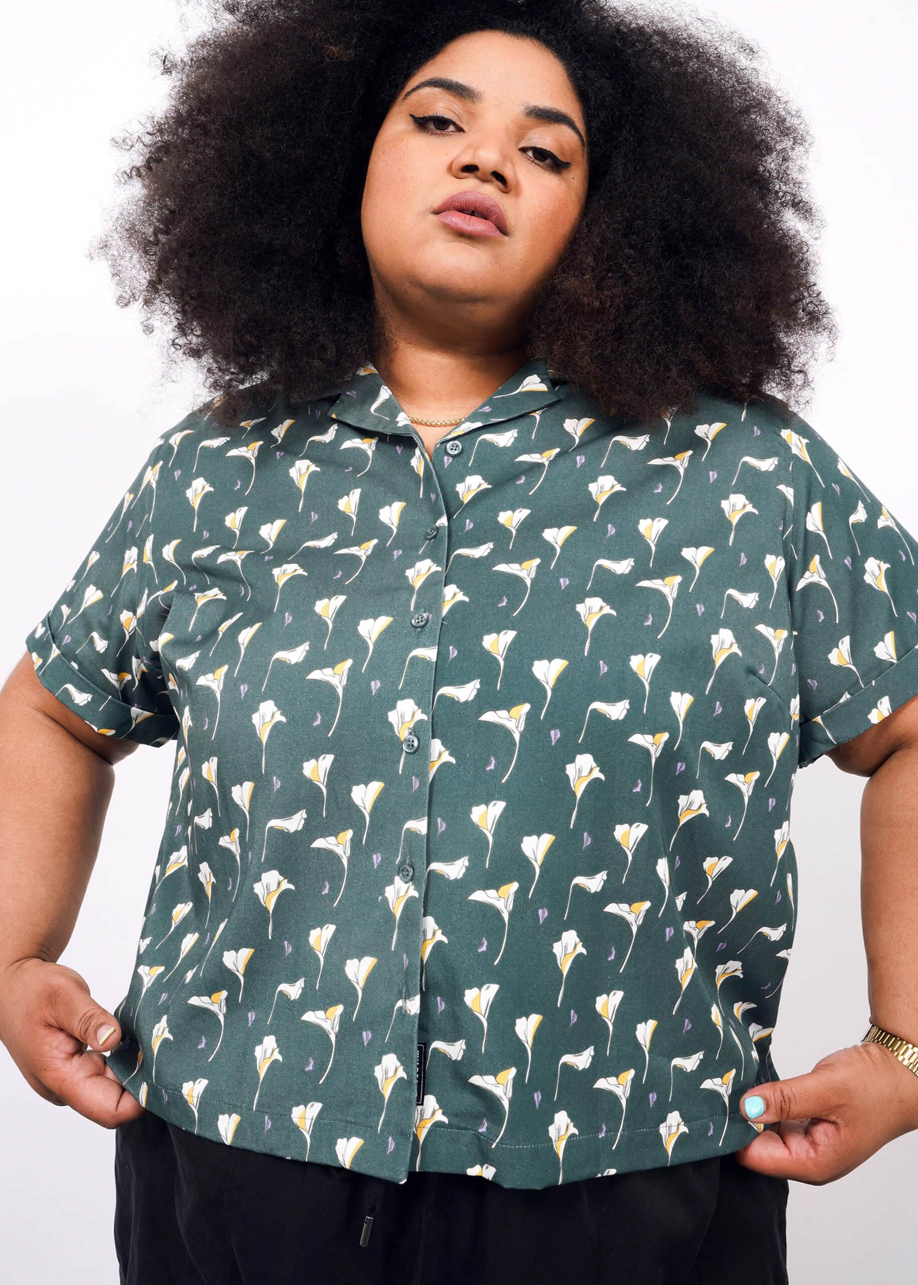 1X= A person with curly hair models The Everyday Camp Button Up in green with white and yellow floral patterns, standing against a plain white background.