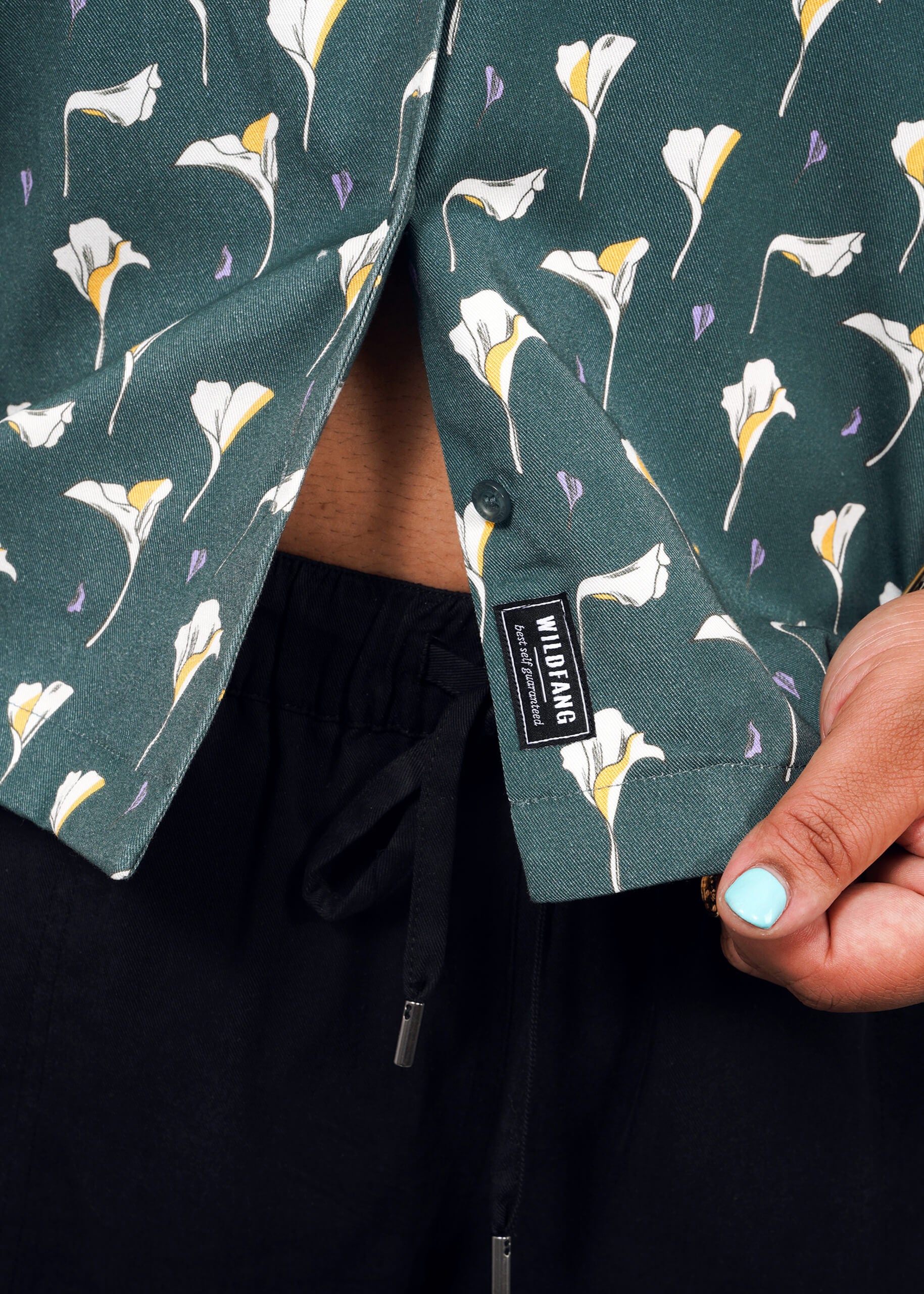 1X=A person opens a green floral-patterned The Everyday Camp Button Up shirt with a visible WILD BANG tag, showing part of their abdomen. They wear black drawstring pants and have mint green nail polish on their thumb.