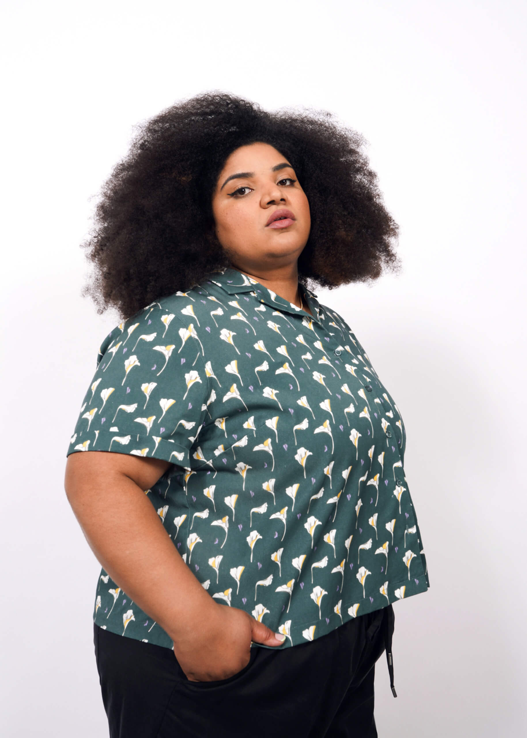1X= A person with curly, voluminous hair wears The Everyday Camp Button Up—a green, short-sleeve camp collar shirt with a floral design and relaxed fit—standing with one hand in their pocket against a plain white background.