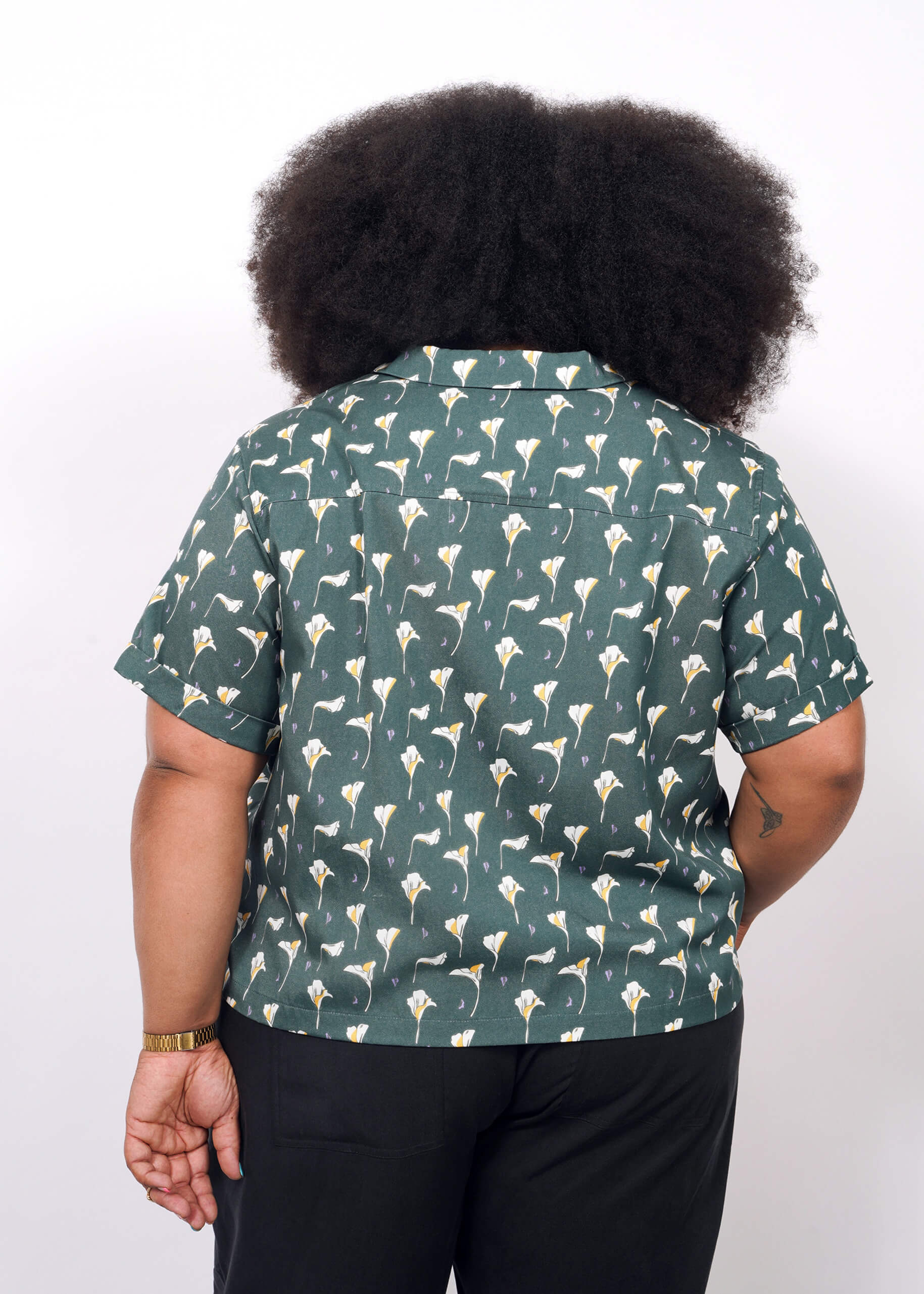 1X=A person with voluminous curly hair is seen from behind wearing The Everyday Camp Button Up—an ultra-soft green shirt with white flowers—and black pants, set against a plain white background.