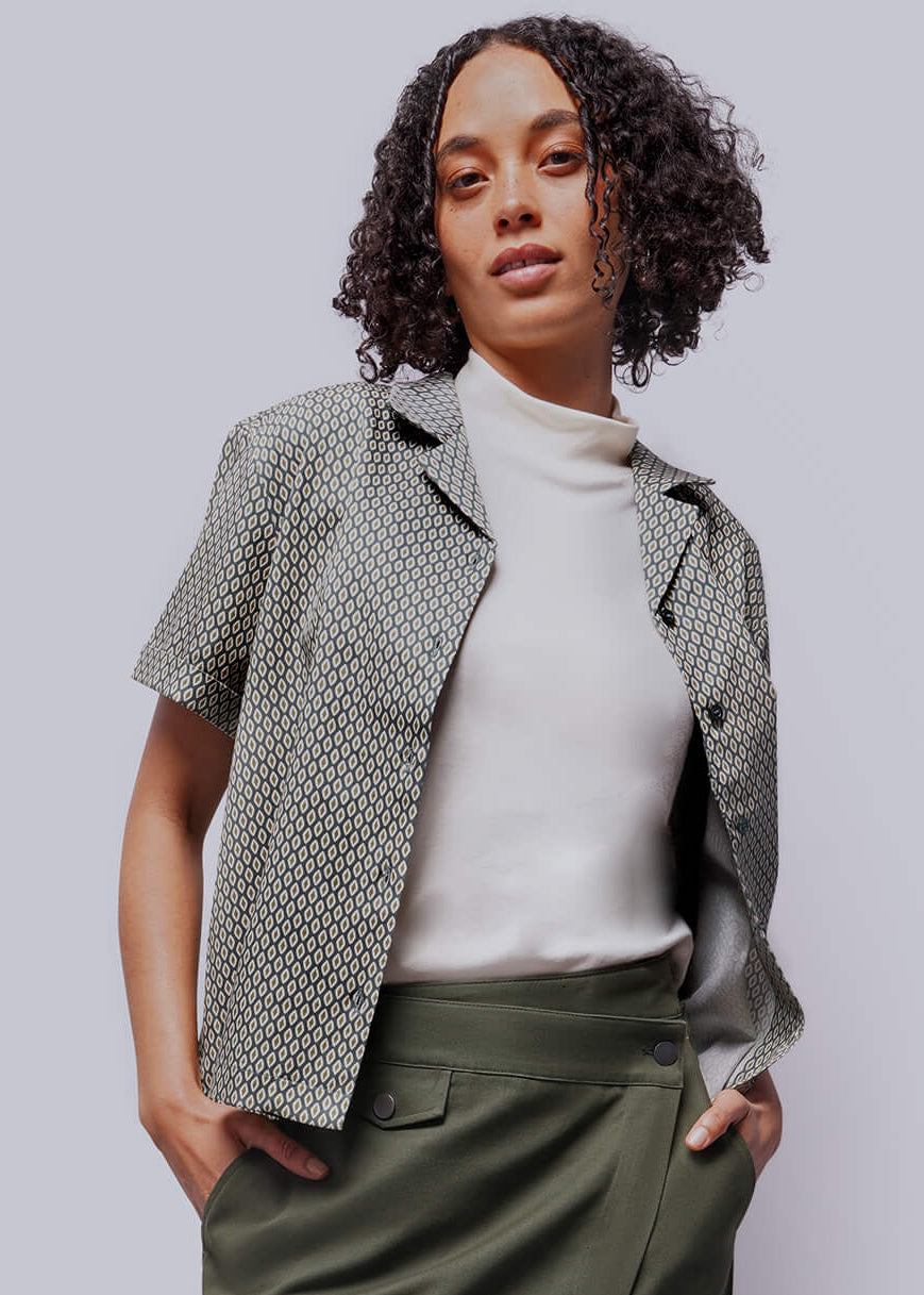 S=Woman wearing a green skirt, white top, and patterned butotn up against a plain background