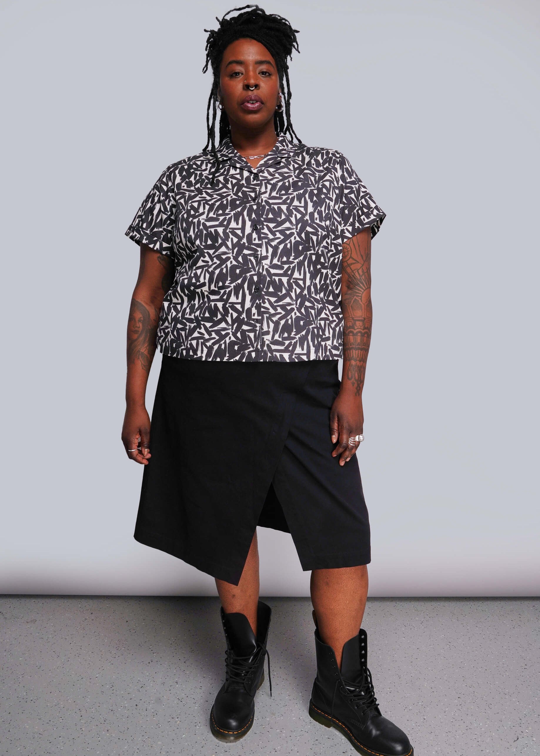 1X=Person wearing a patterned shirt and black skirt with black boots against a plain background