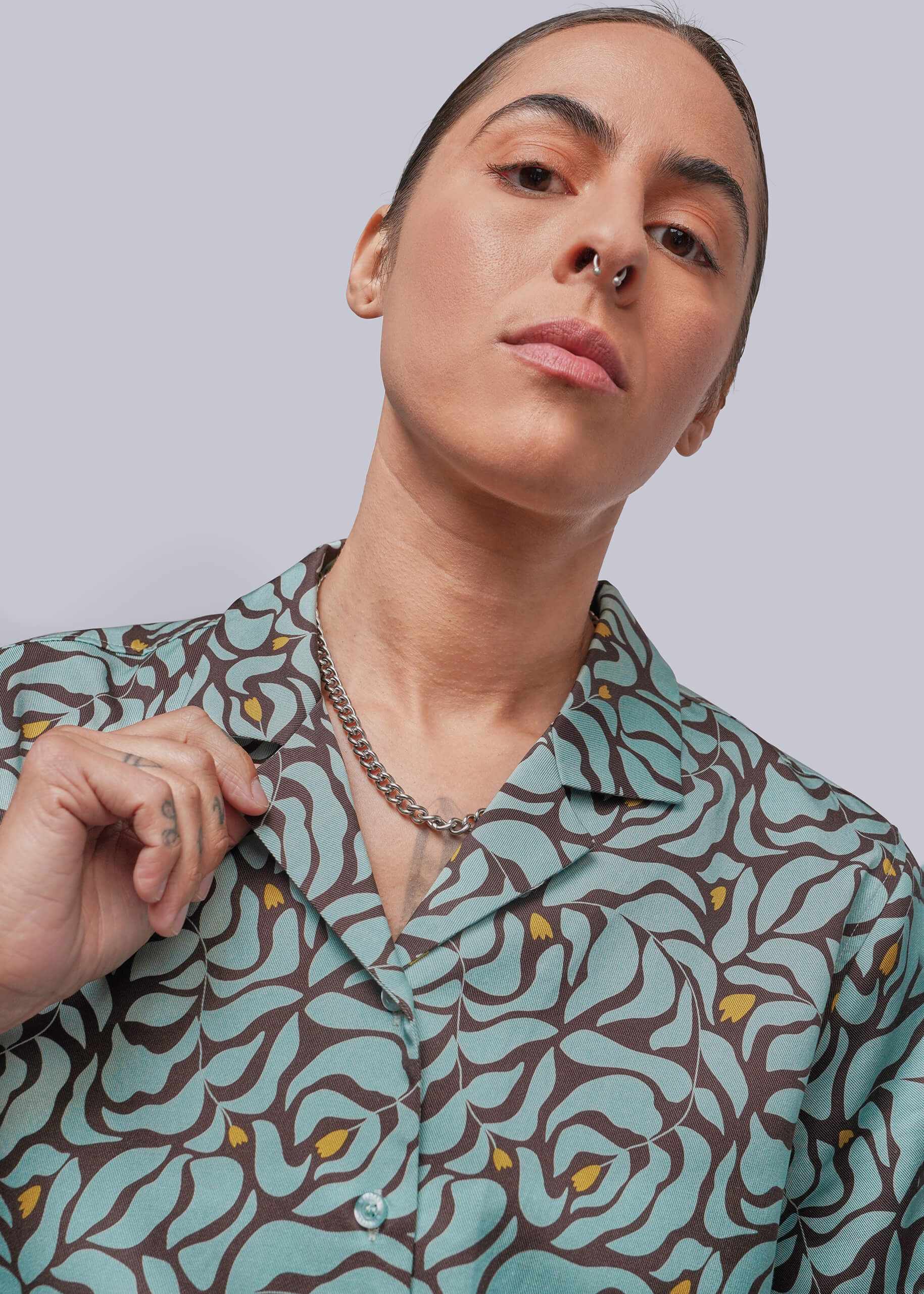 S=A close-up of a model wearing a collared button-up shirt with a brown and mint green abstract wavy pattern and small yellow accents, paired with a silver chain necklace.