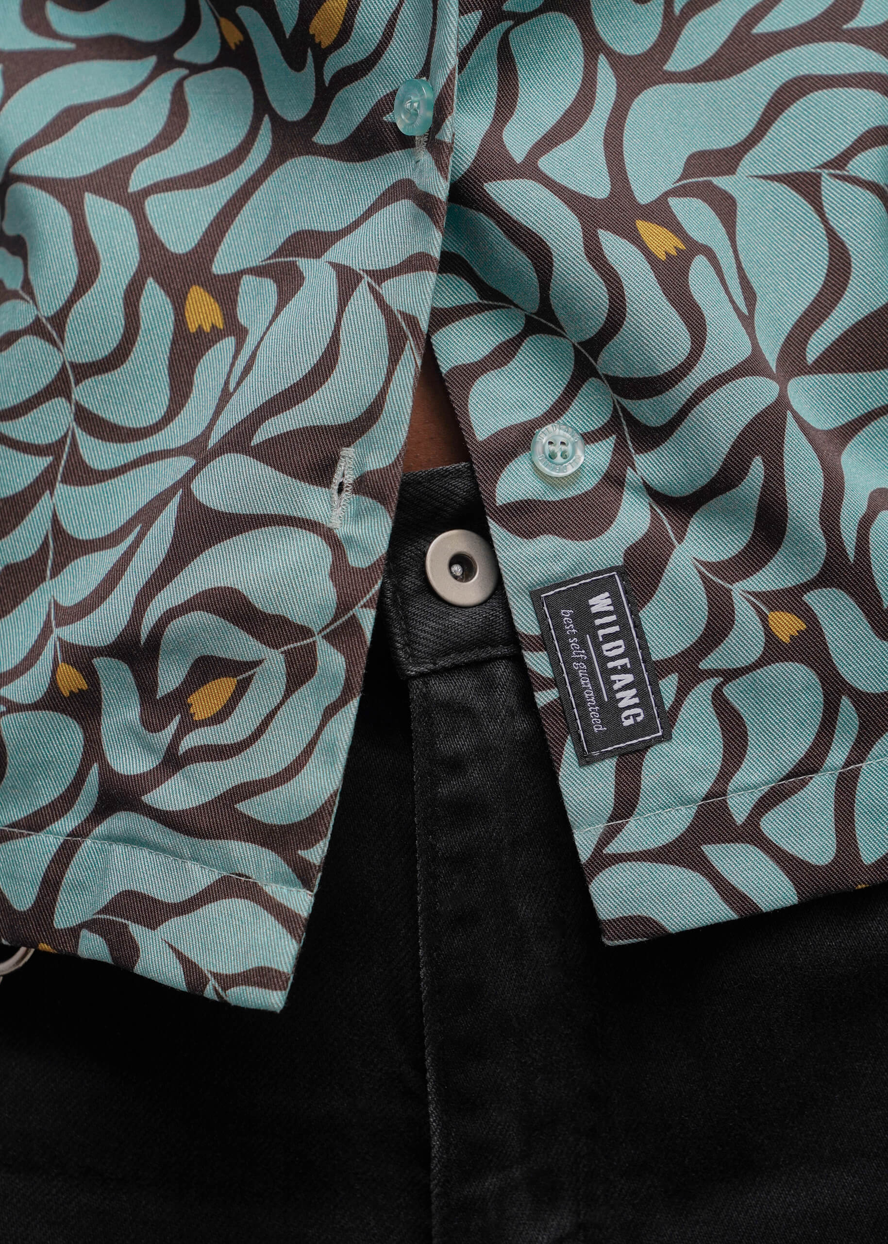 S=Close-up of a button-front shirt with an abstract wavy pattern in light aqua and dark brown, with small mustard-yellow accents. A black fabric brand tab reading WILDFANG is visible near the placket, and the shirt is styled over a black garment.