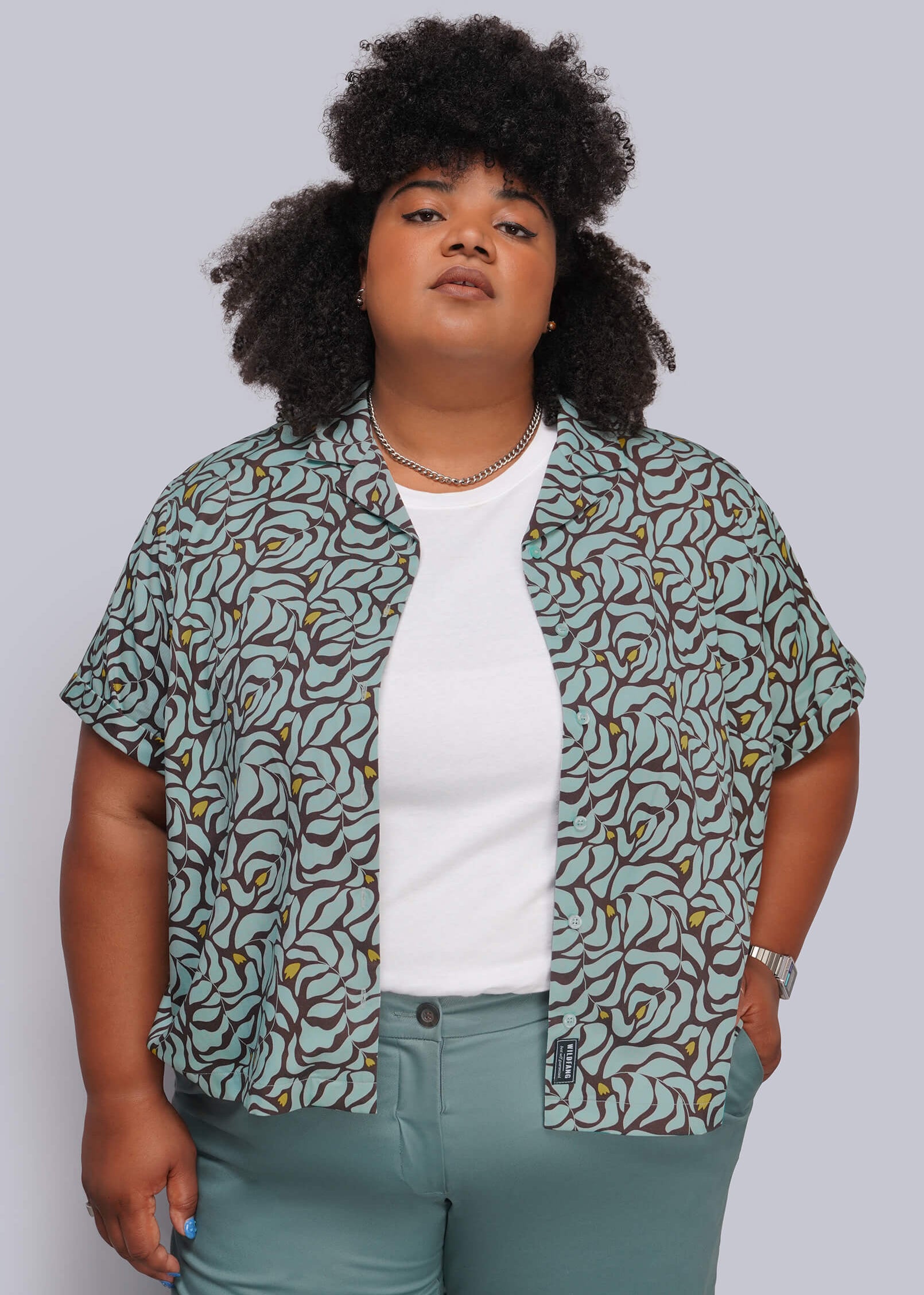 1X=A plus-size model wearing a short-sleeve button-up shirt with a green, brown, and beige abstract pattern over a white t-shirt and sage green pants, set against a solid gray background.