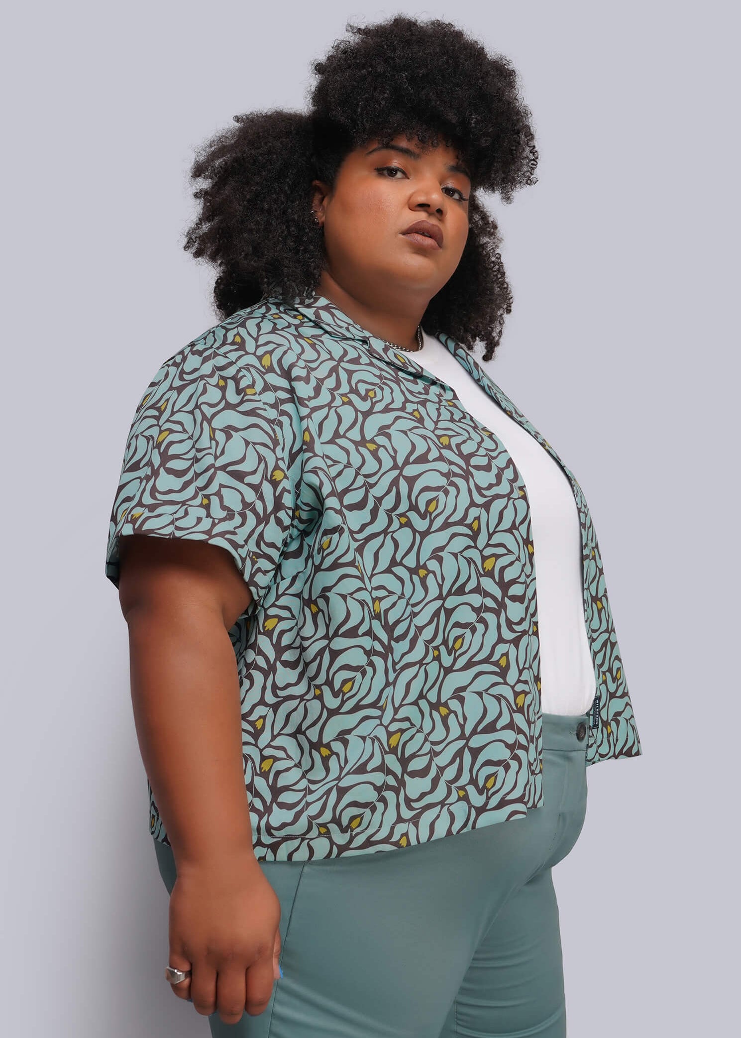 1X=A woman modeling a short-sleeve button-up shirt with a green, brown, and yellow abstract pattern, worn open over a white top and paired with teal pants, against a solid gray studio background.