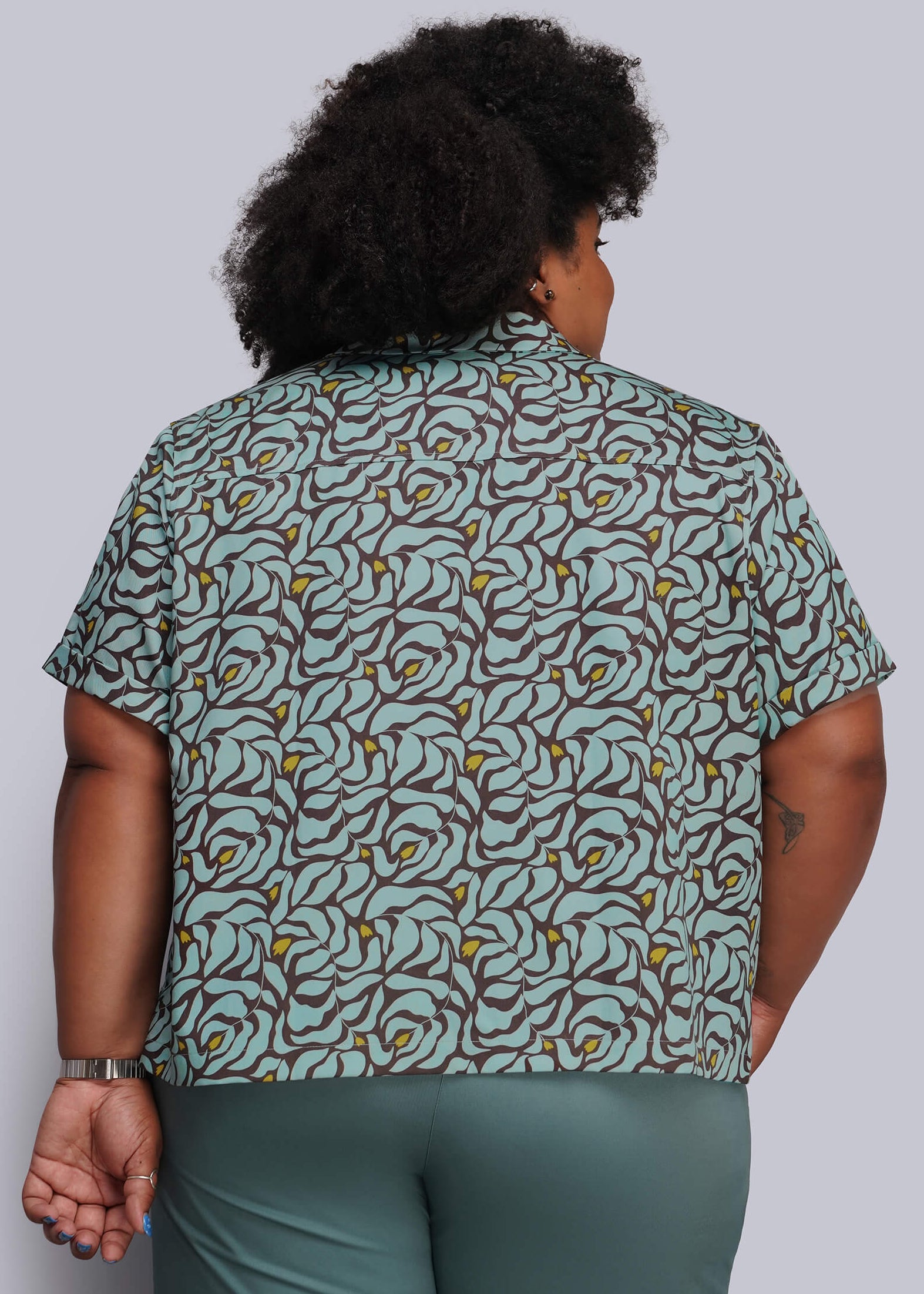 1X=A person shown from the back wearing a short-sleeve collared button-up shirt with an abstract floral or leaf-like print in muted green, brown, and yellow, paired with solid green pants, against a plain light gray studio background.