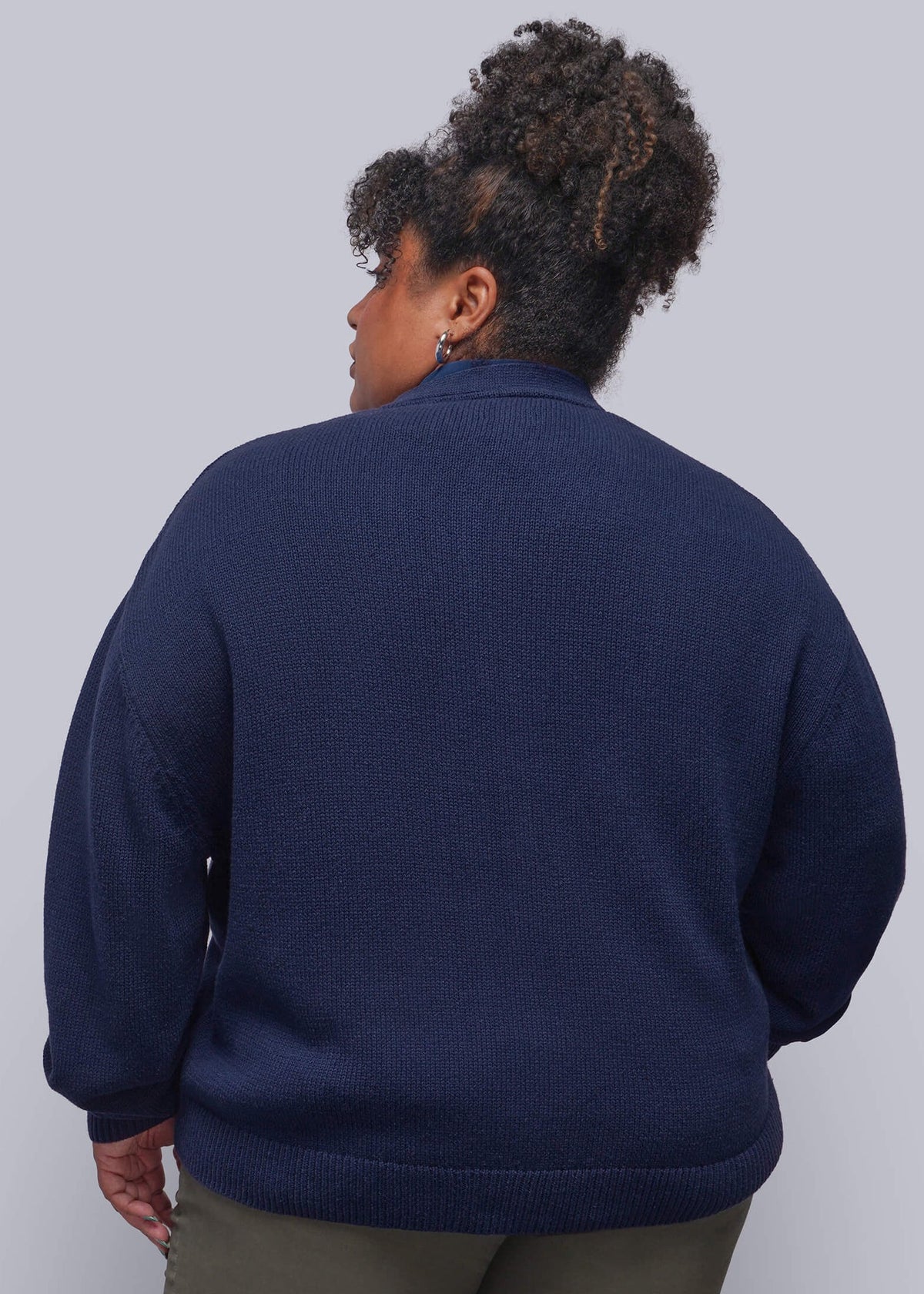 1X=A person shown from the back wearing a navy blue knit sweater and olive green pants against a light gray studio background.