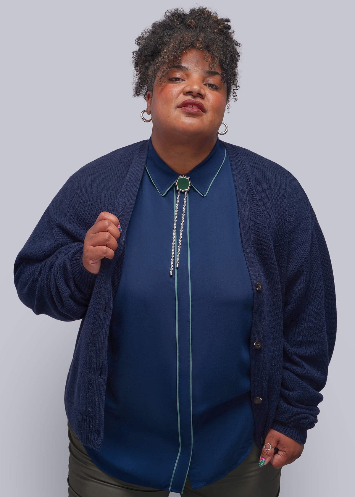1X=A plus-size model wearing a navy blue cardigan over a matching navy blue collared blouse with a beaded bolo tie, paired with olive green pants, against a solid gray studio background.
