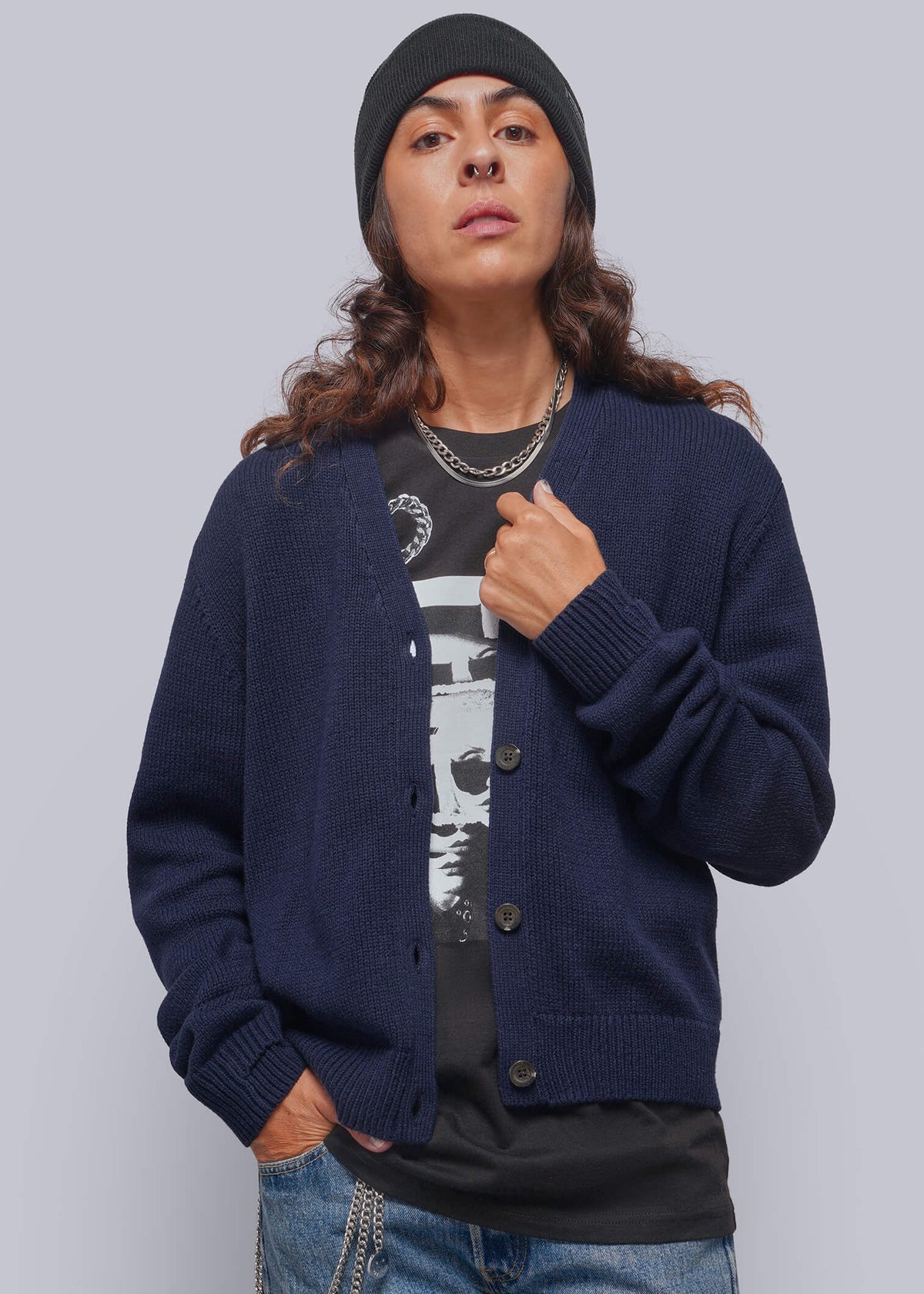 S=A model wearing a navy blue knit cardigan over a black graphic t-shirt, paired with blue jeans, a black beanie, and silver chain accessories, set against a solid gray studio background.