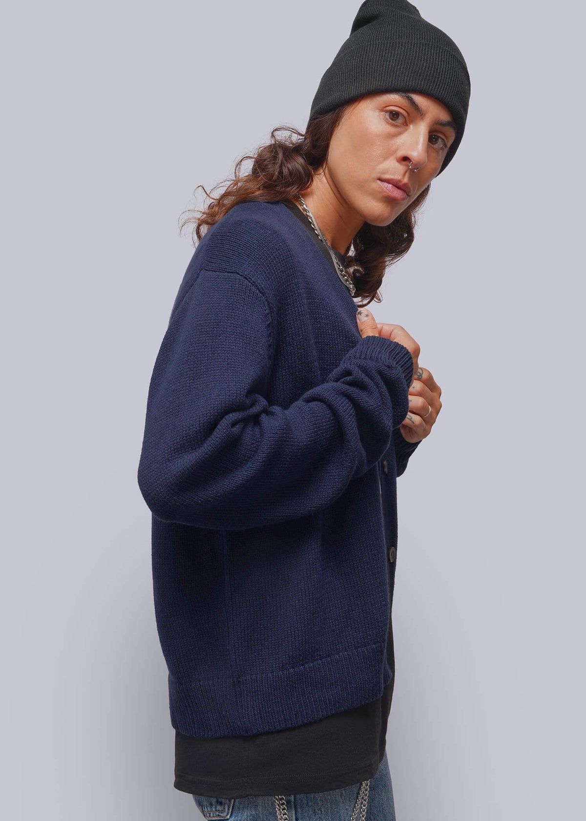 S=A person wearing a navy blue knit sweater, a black beanie, and blue jeans with silver chain details, photographed against a solid light gray studio background.