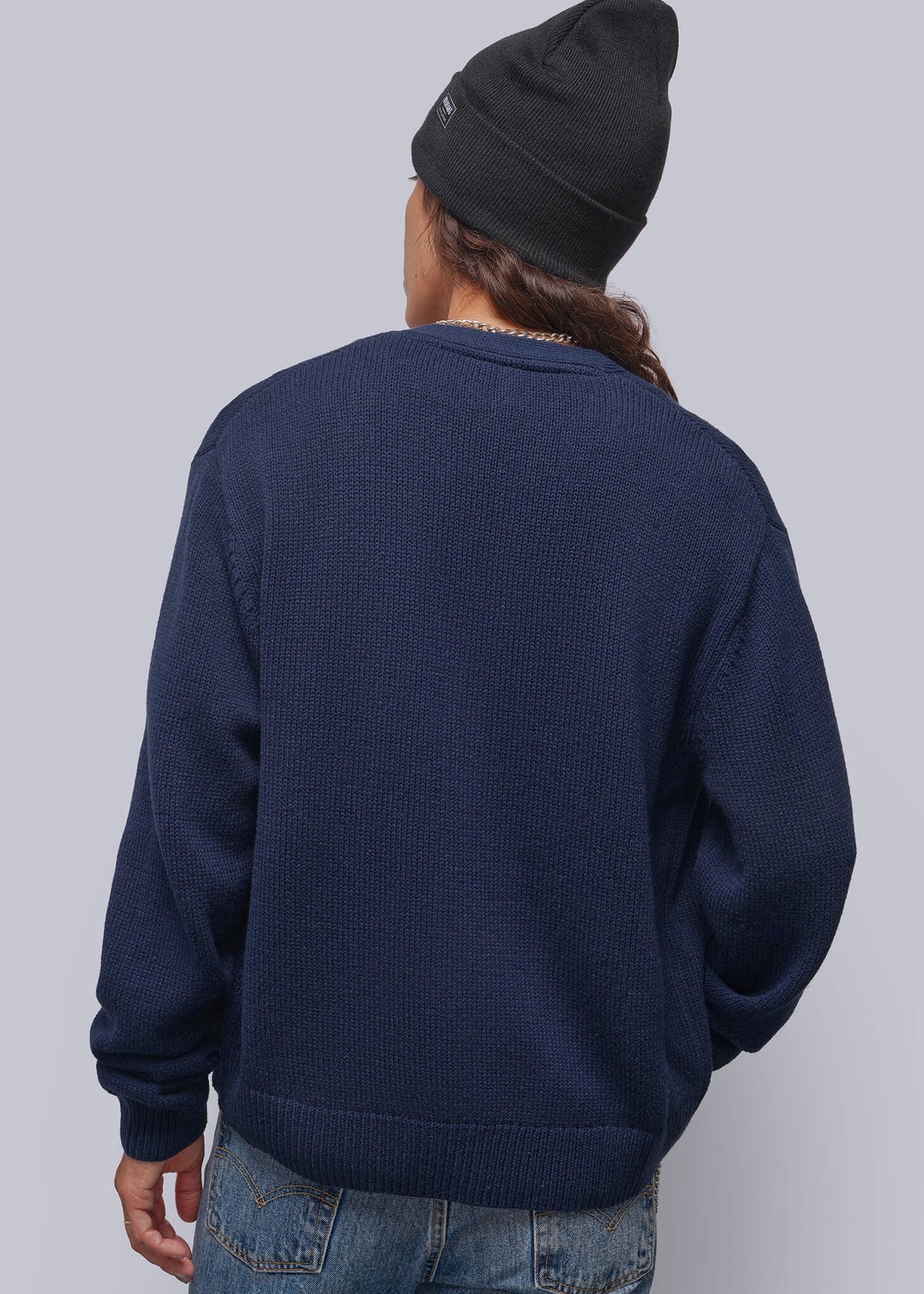 S=A person shown from the back wearing a navy blue knit sweater, blue jeans, and a black beanie against a light gray studio background.