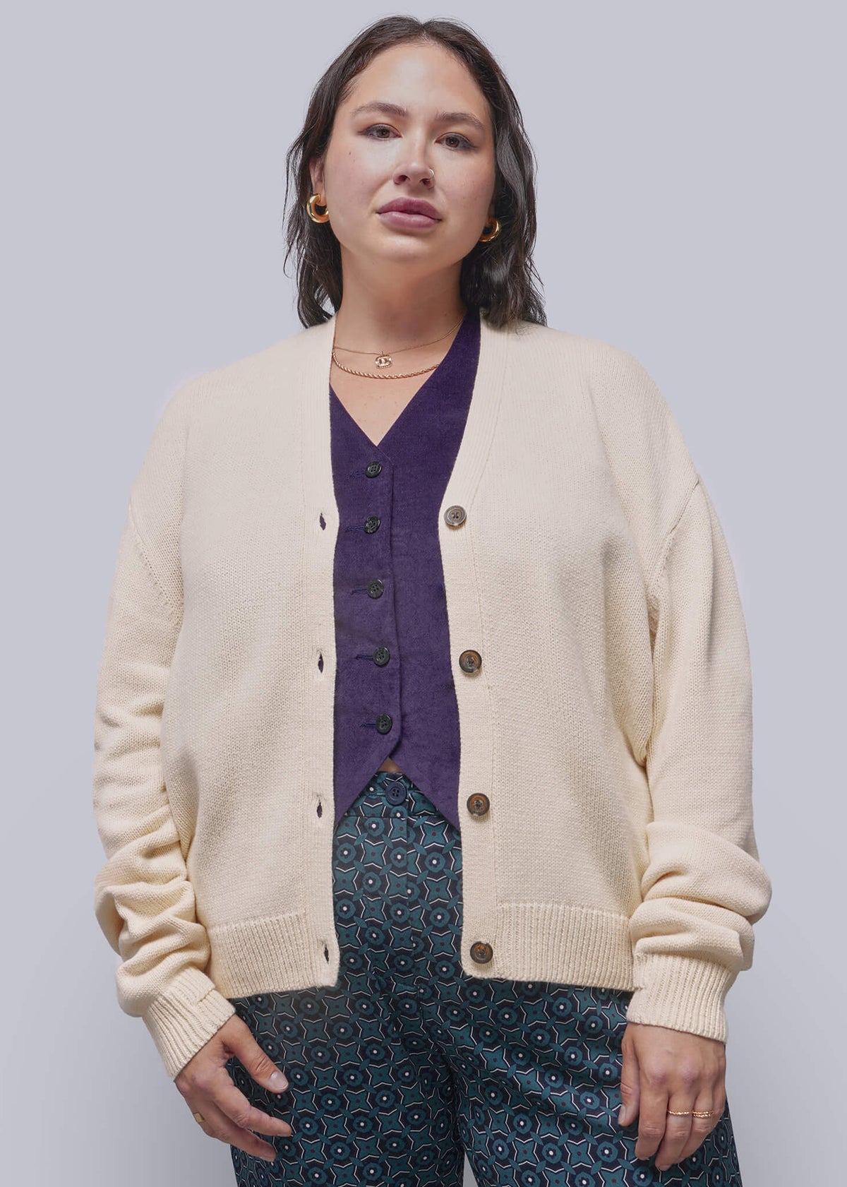 1X=A woman modeling a cream-colored knit cardigan over a dark purple V-neck top and dark blue patterned pants, shot against a solid light gray studio background.