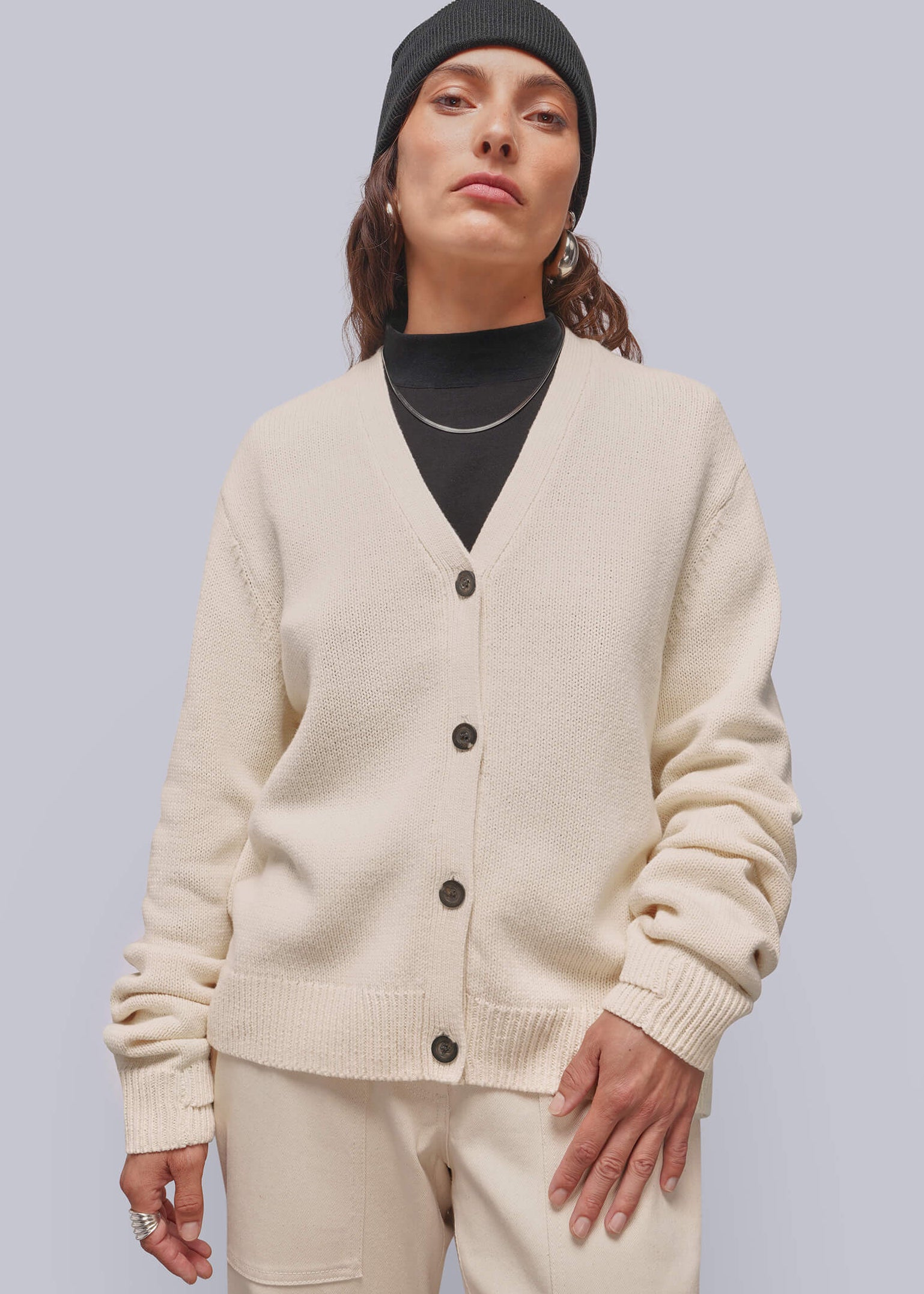 A model wearing a cream-colored knit cardigan and matching trousers, styled with a black turtleneck, black beanie, and silver necklace against a solid light gray background.