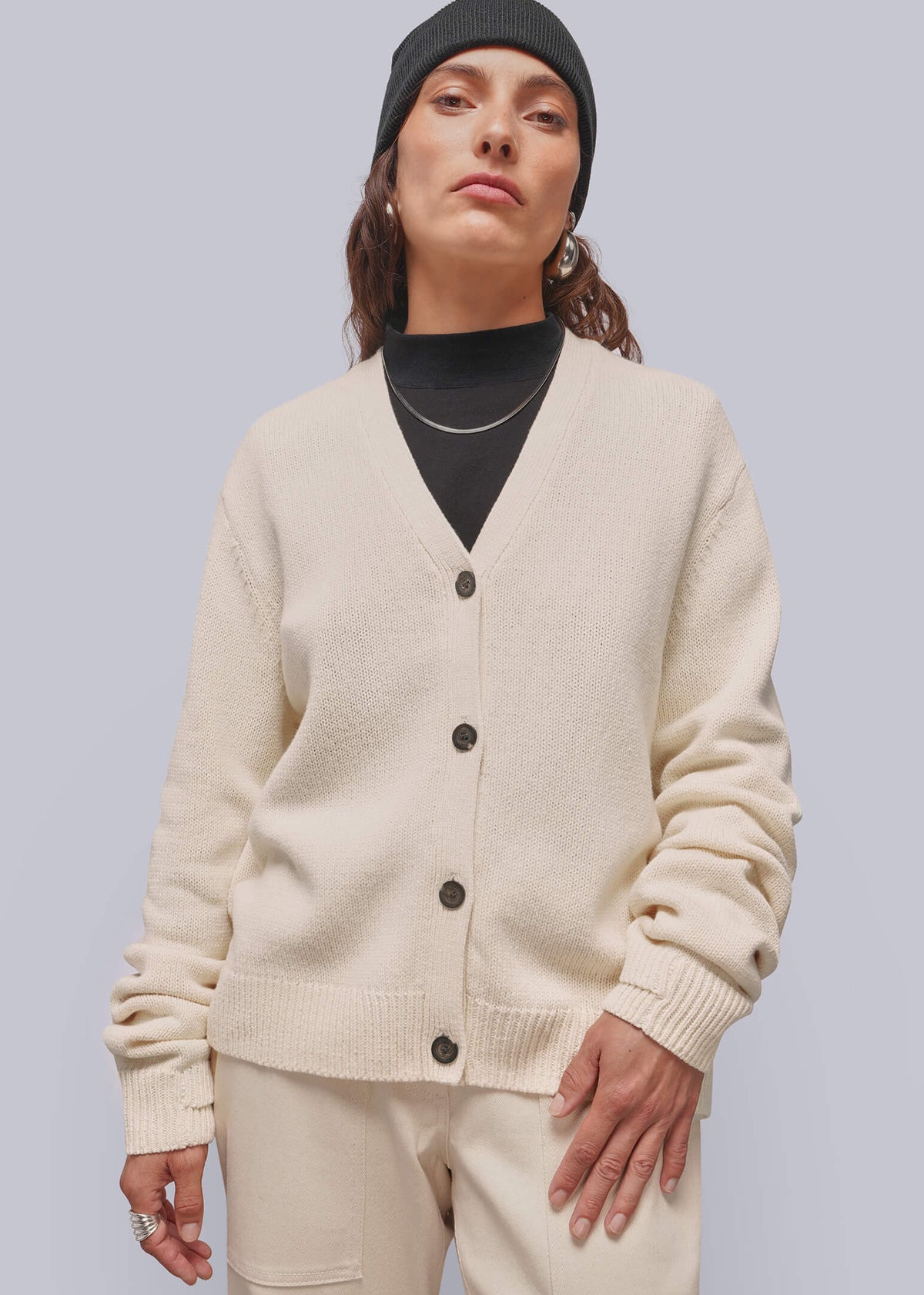 S=A model wearing a cream-colored knit cardigan and matching trousers, styled with a black turtleneck, black beanie, and silver necklace against a solid light gray background.