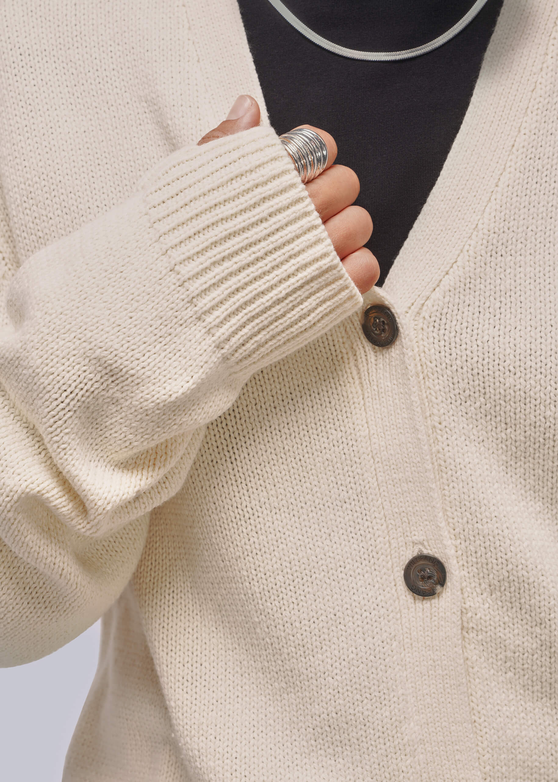 Close-up of a cream-colored knit cardigan sweater with dark brown buttons, worn over a black top. A hand with a silver ring is shown adjusting the ribbed cuff of the cardigan.