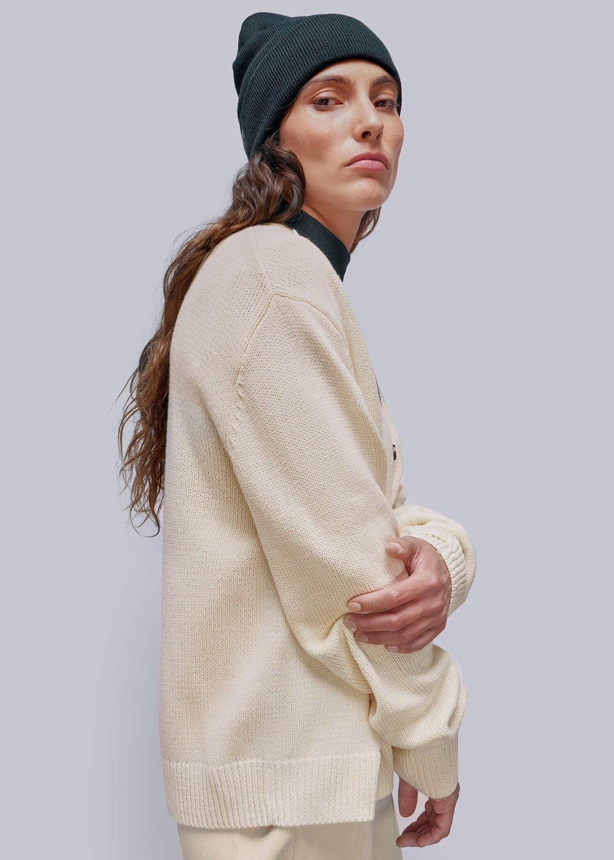 S=A woman wearing a cream-colored knit cardigan, matching cream pants, a dark green beanie, and a black collar shirt, photographed against a solid light gray background.