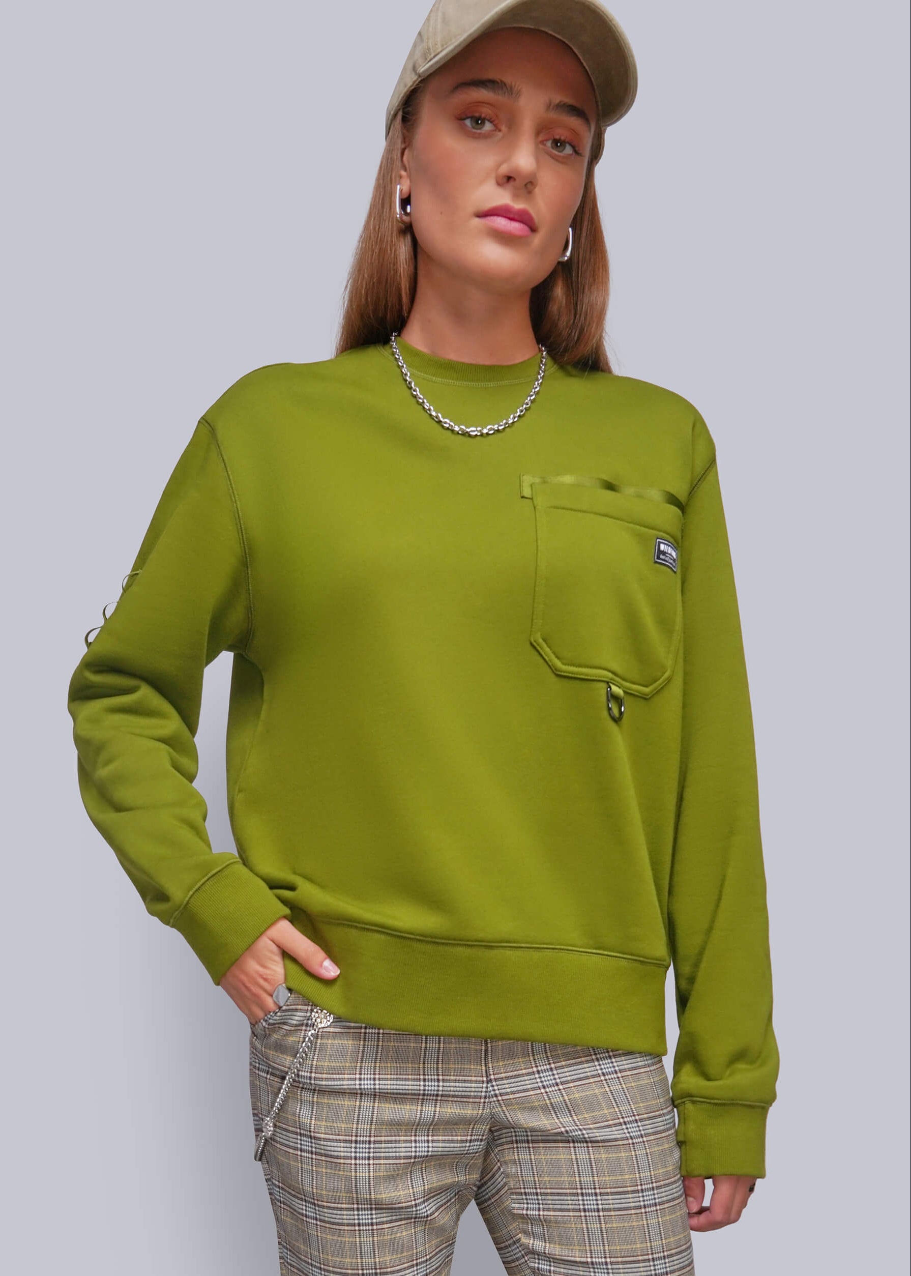 A woman wearing a green crewneck sweatshirt with a chest pocket, paired with beige plaid pants and a beige cap, against a solid gray studio background.