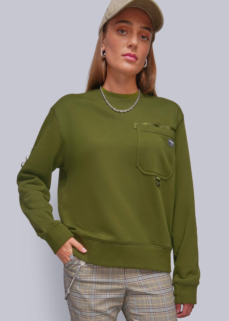 S=A woman wearing a green crewneck sweatshirt with a chest pocket, paired with beige plaid pants and a beige cap, against a solid gray studio background.
