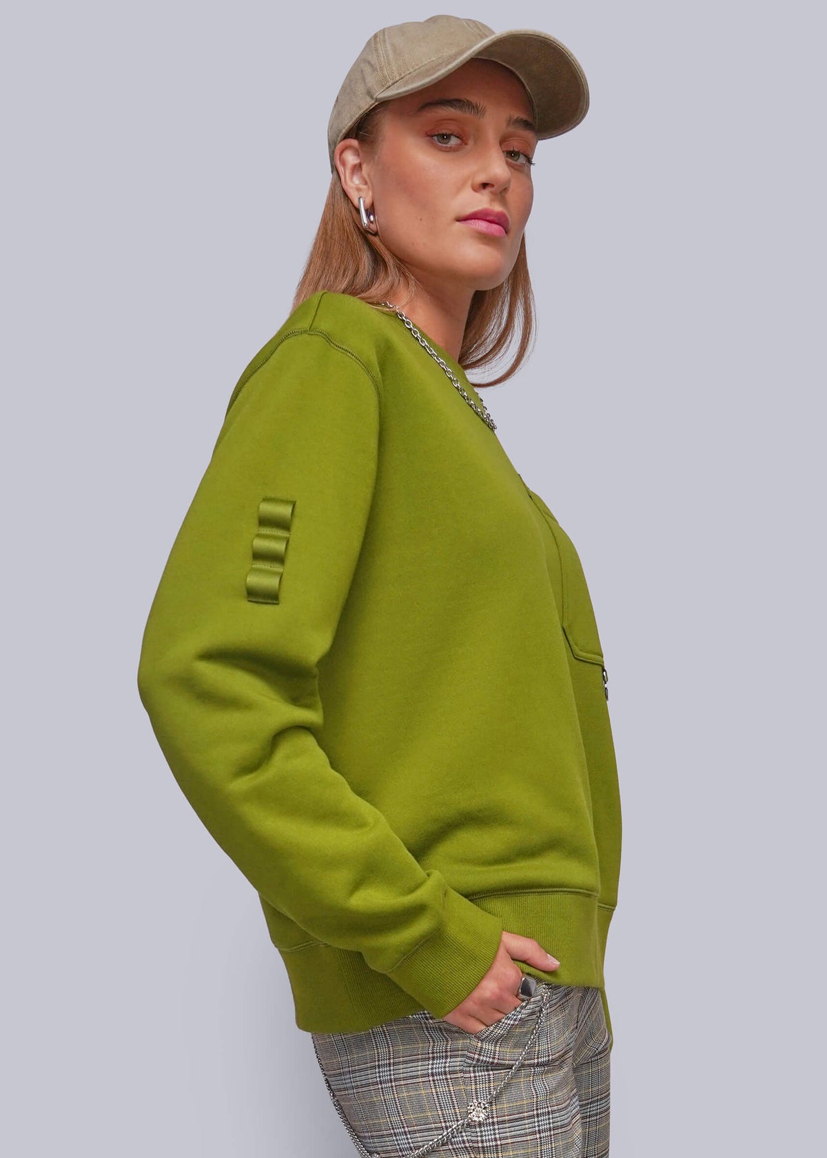 S=A woman wearing a green crewneck sweatshirt with unique ribbed sleeve detailing, paired with plaid pants and a beige cap, shown against a solid gray background.