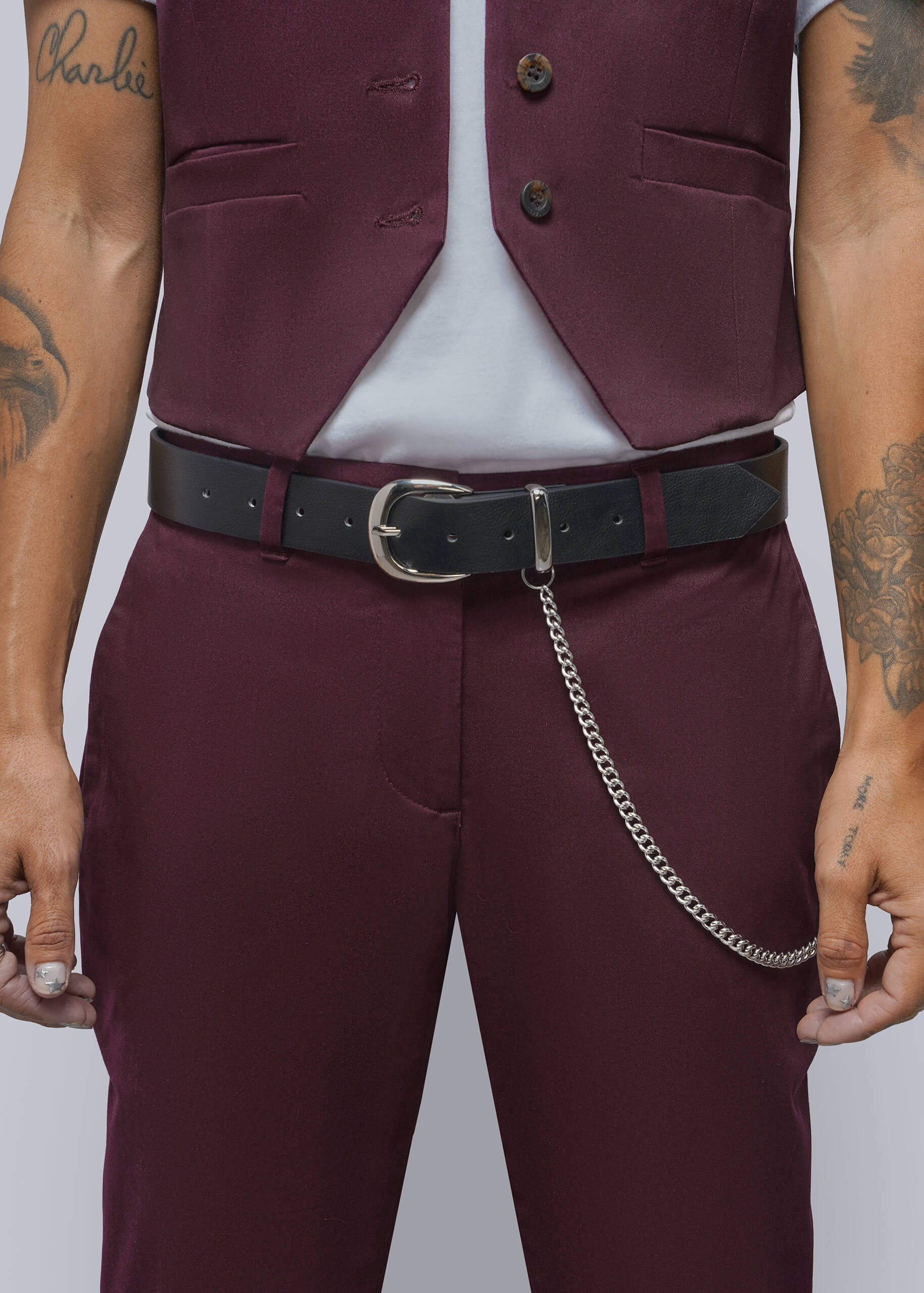 A close-up of a person wearing a tailored outfit featuring a maroon vest and matching maroon trousers, styled with a black belt and silver chain detail. The person has visible tattoos on both arms, and the background is a solid light gray.