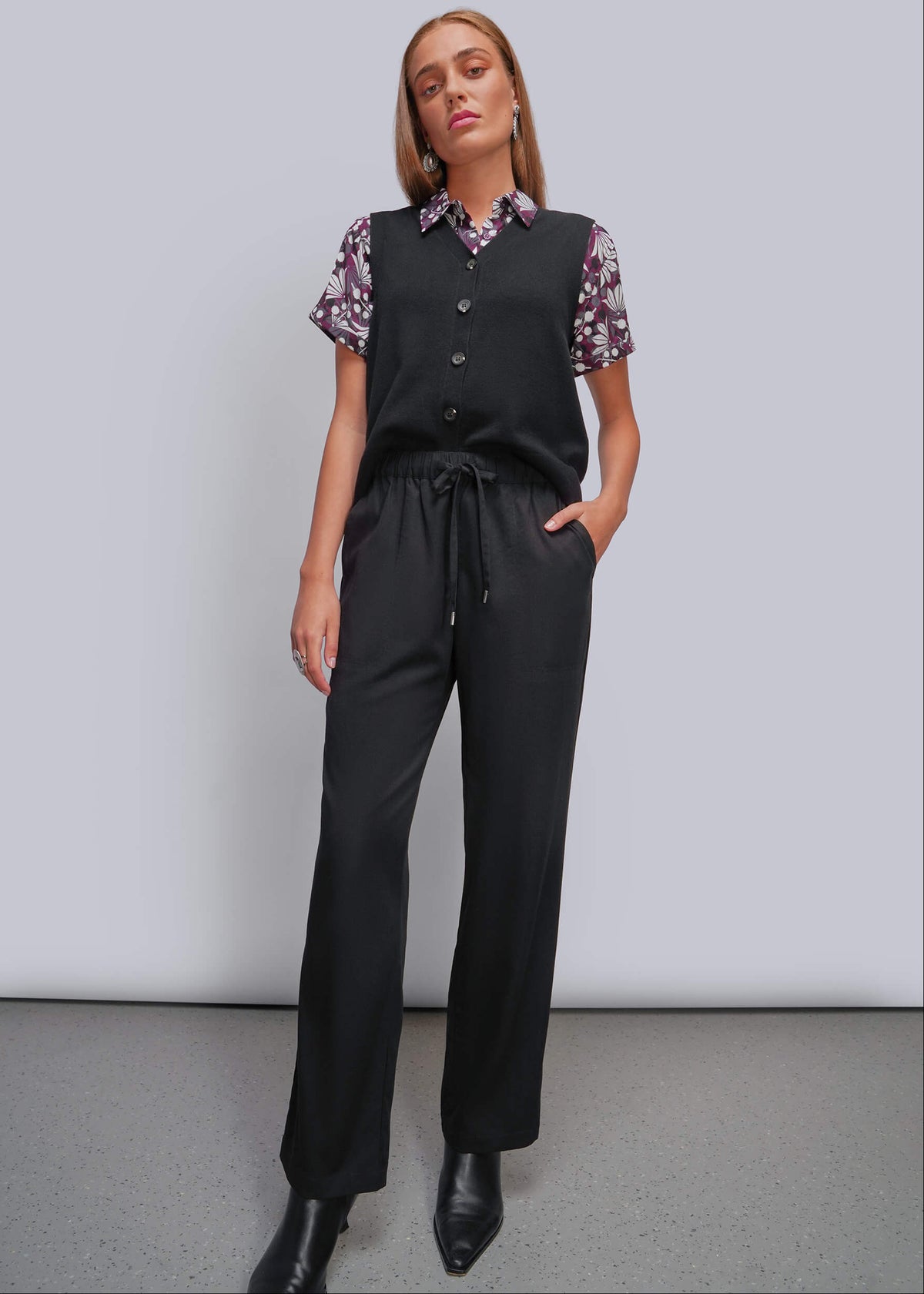 S=A woman modeling a black sleeveless vest and matching black drawstring trousers over a short-sleeve floral collared shirt, standing against a light gray studio background.