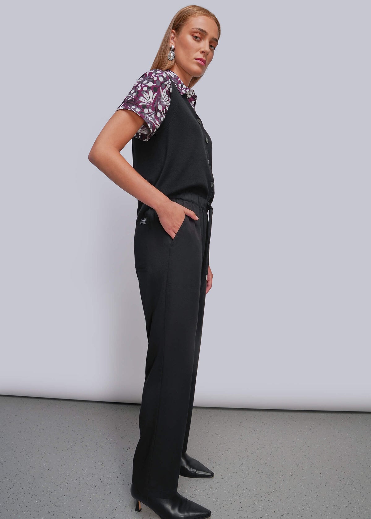 S=A woman models a black sleeveless vest and matching wide-leg trousers, styled with a short-sleeve floral shirt and black pointed-toe heels against a light gray studio background.