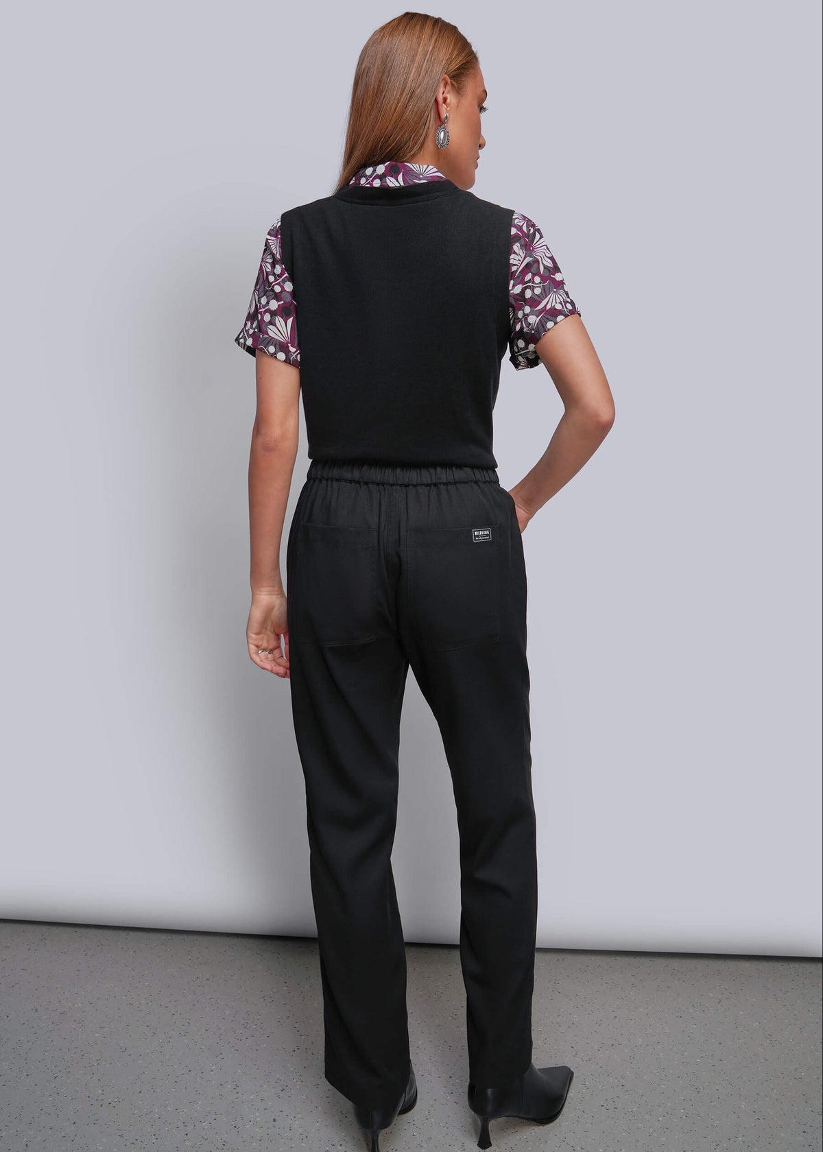 S=A woman is shown from behind wearing a black sleeveless vest over a short-sleeve floral shirt and black trousers, standing against a light gray studio wall.