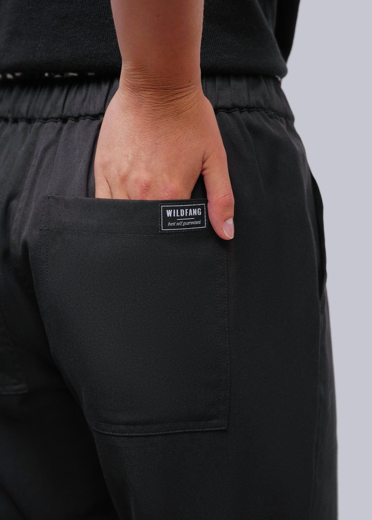 S=Close-up of a person wearing black pants, with a hand in the back pocket showing a small black brand label that reads WILDFANG.