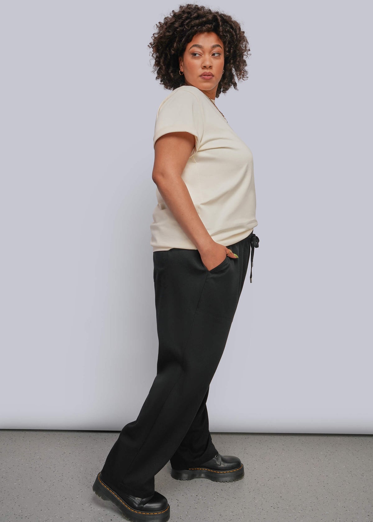 1X=Side view of model wearing off white tshirt with black drawstring pant and black boots
