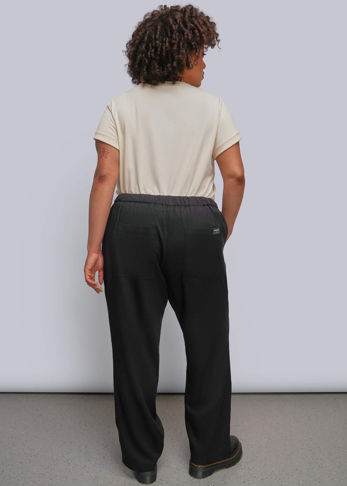 1X=A person shown from the back wearing loose black pants and a light beige short-sleeve top, standing against a plain light gray studio wall.