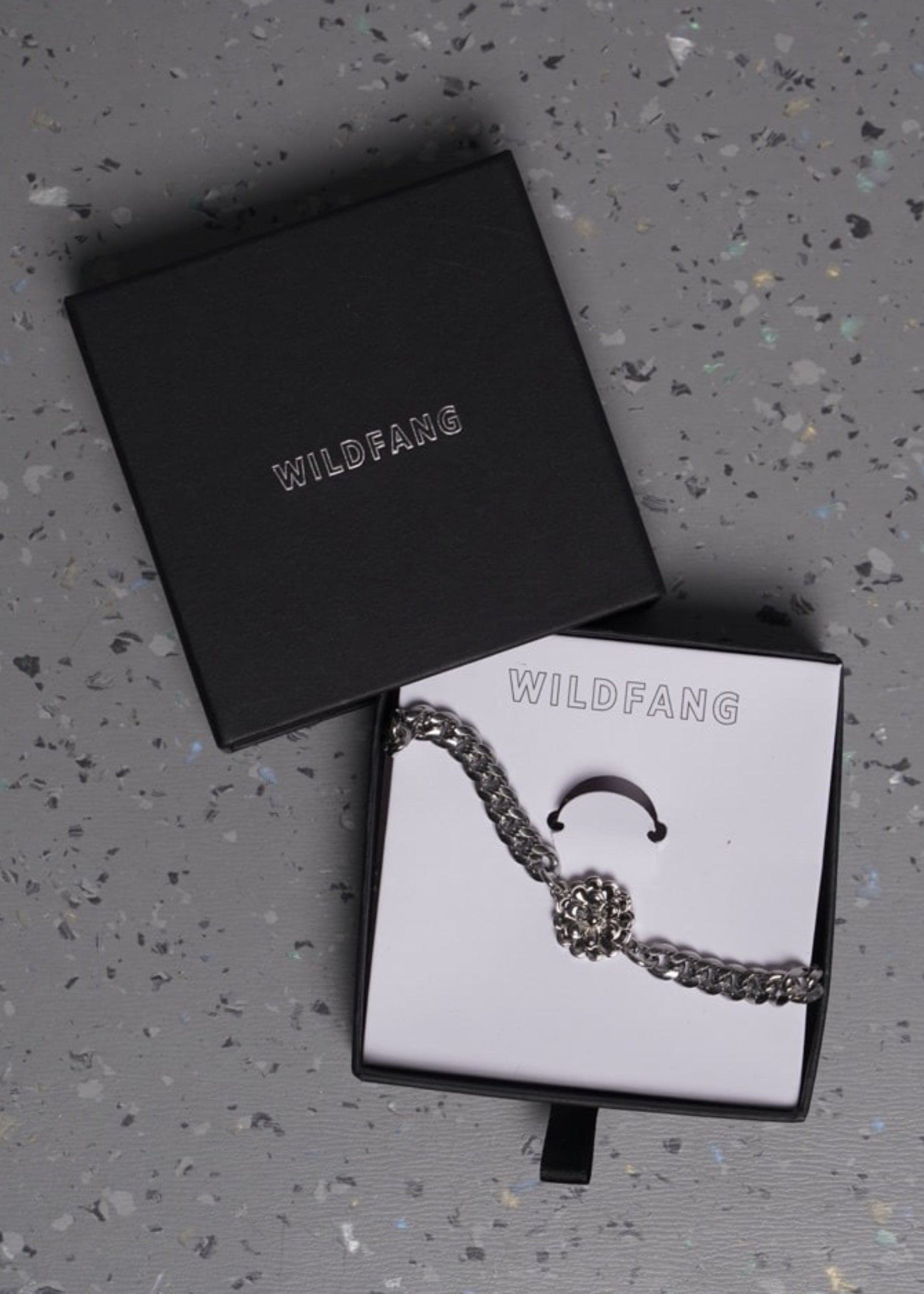 A silver-toned wallet chain with a floral charm, displayed in an open box with the brand name WILDFANG, next to the box lid on a speckled gray surface.