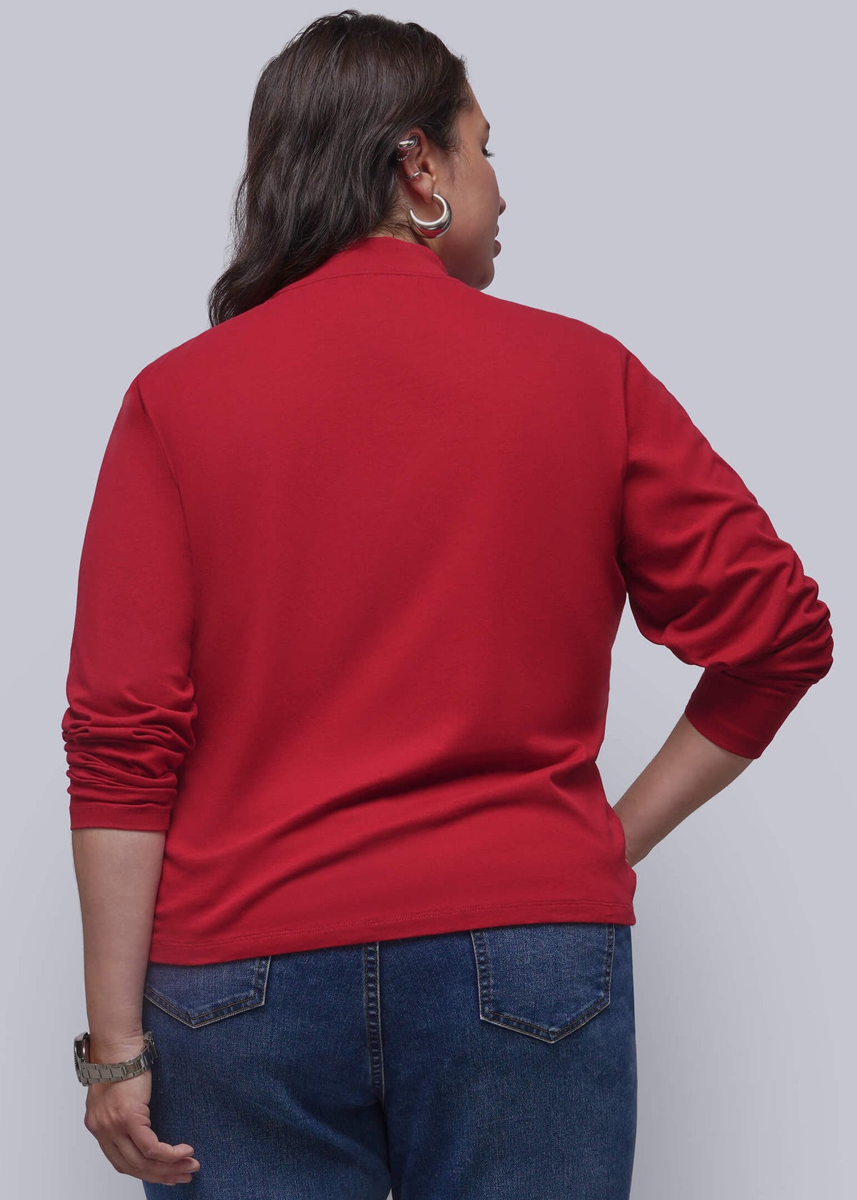 1X=A woman modeling a red mock neck long-sleeve top with blue jeans, shown from the back, standing against a solid gray studio background.