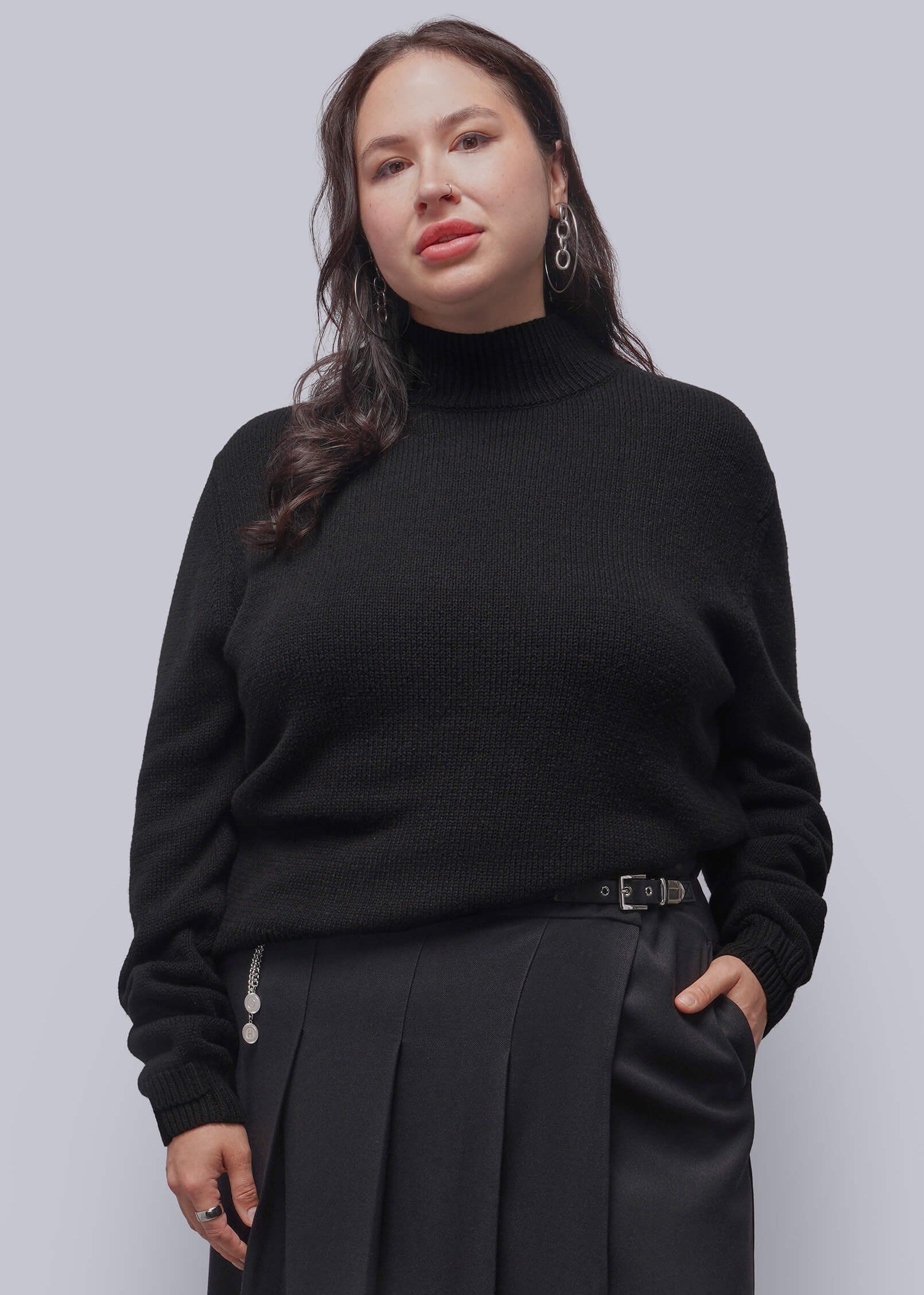 1X=A woman modeling a black ribbed turtleneck sweater and black pleated skirt against a solid gray studio background.