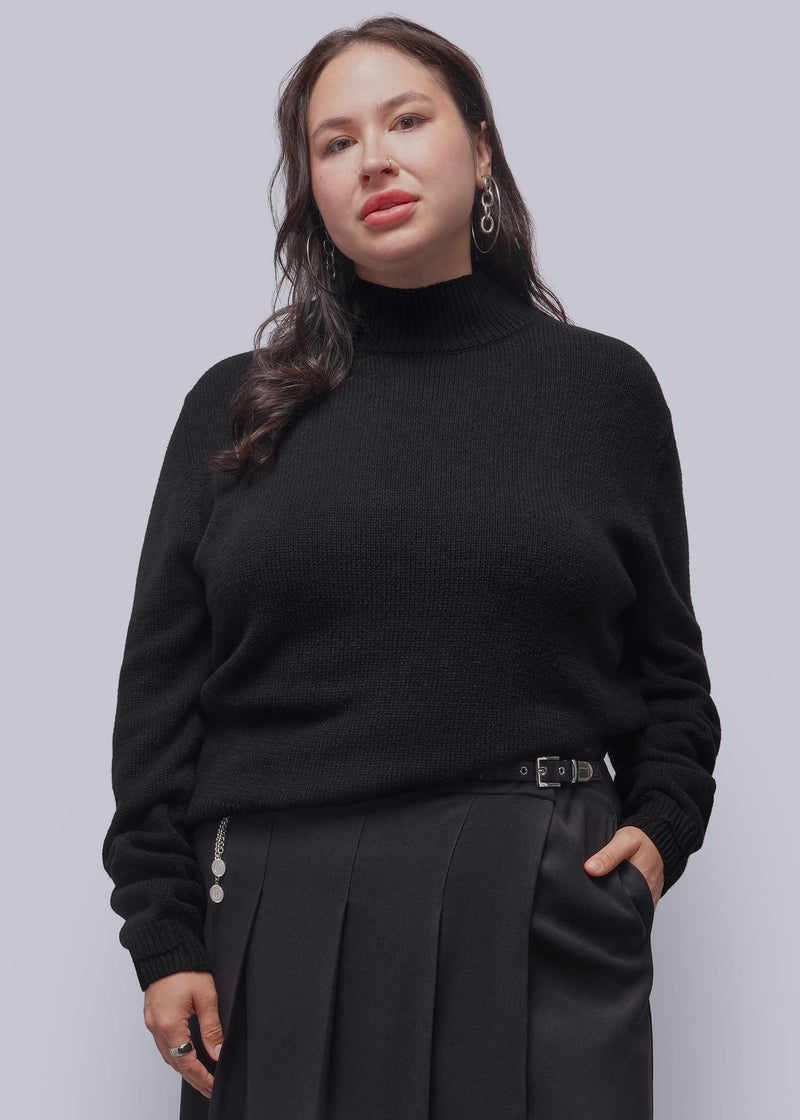 1X=A woman modeling a black ribbed turtleneck sweater and black pleated skirt against a solid gray studio background.
