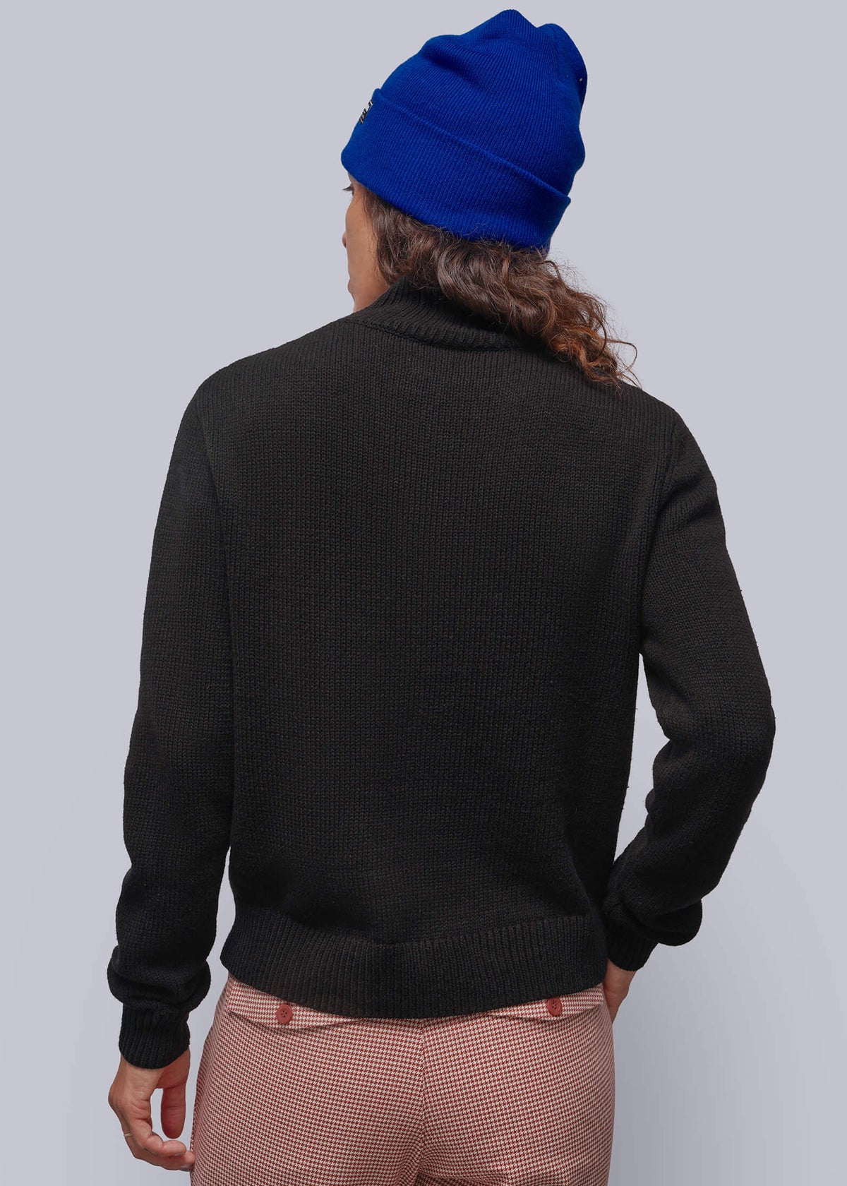 S=A person shown from the back wearing a black knit sweater, red houndstooth patterned trousers, and a bright blue beanie against a light gray studio background.