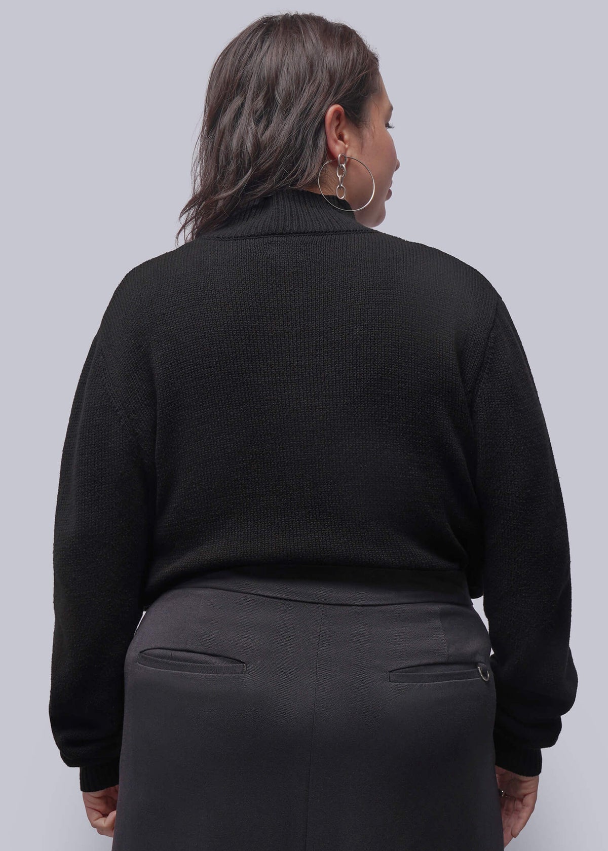 1X=A woman shown from the back wearing a black ribbed turtleneck sweater and a dark gray skirt, photographed against a solid light gray studio background.