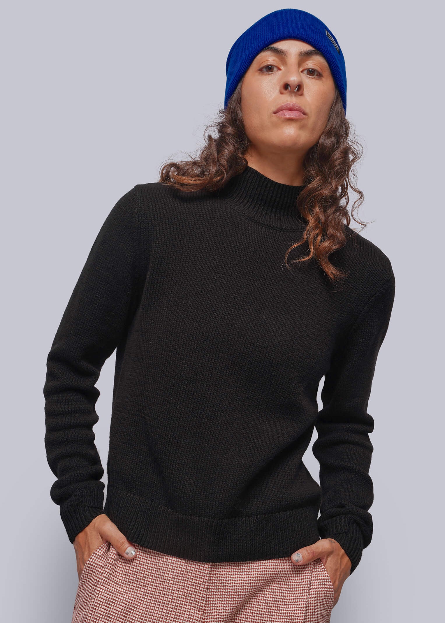 A person wearing a black mock neck knit sweater, red patterned houndstooth trousers, and a bright blue beanie, standing against a solid gray studio background.