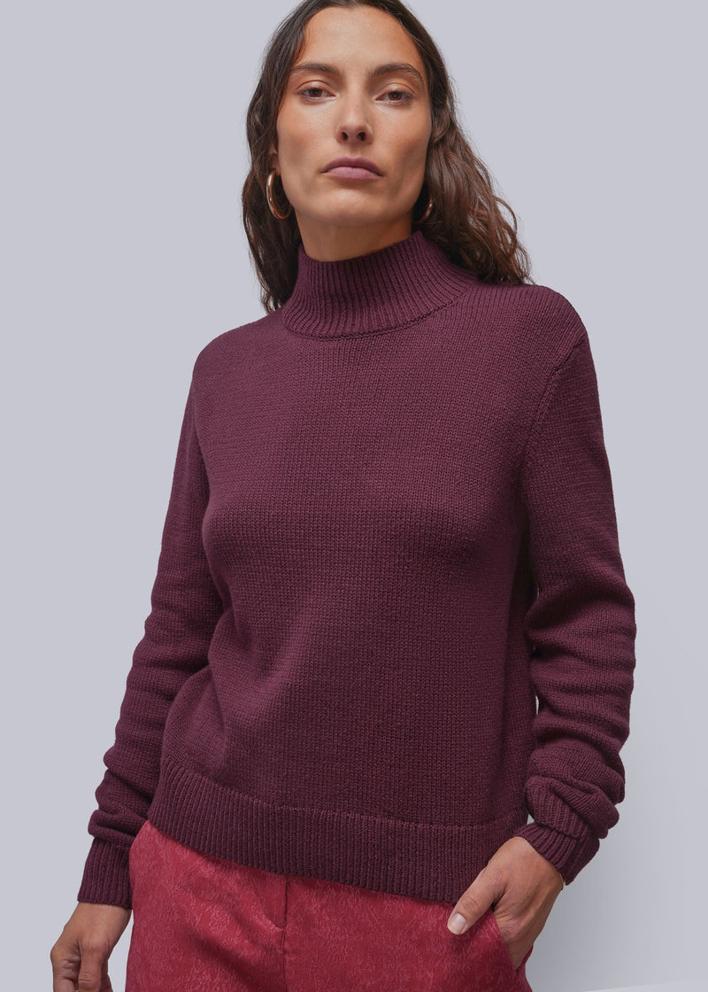 S=A woman wearing a ribbed turtleneck sweater in a deep burgundy color, paired with textured pink pants, against a solid light gray background.