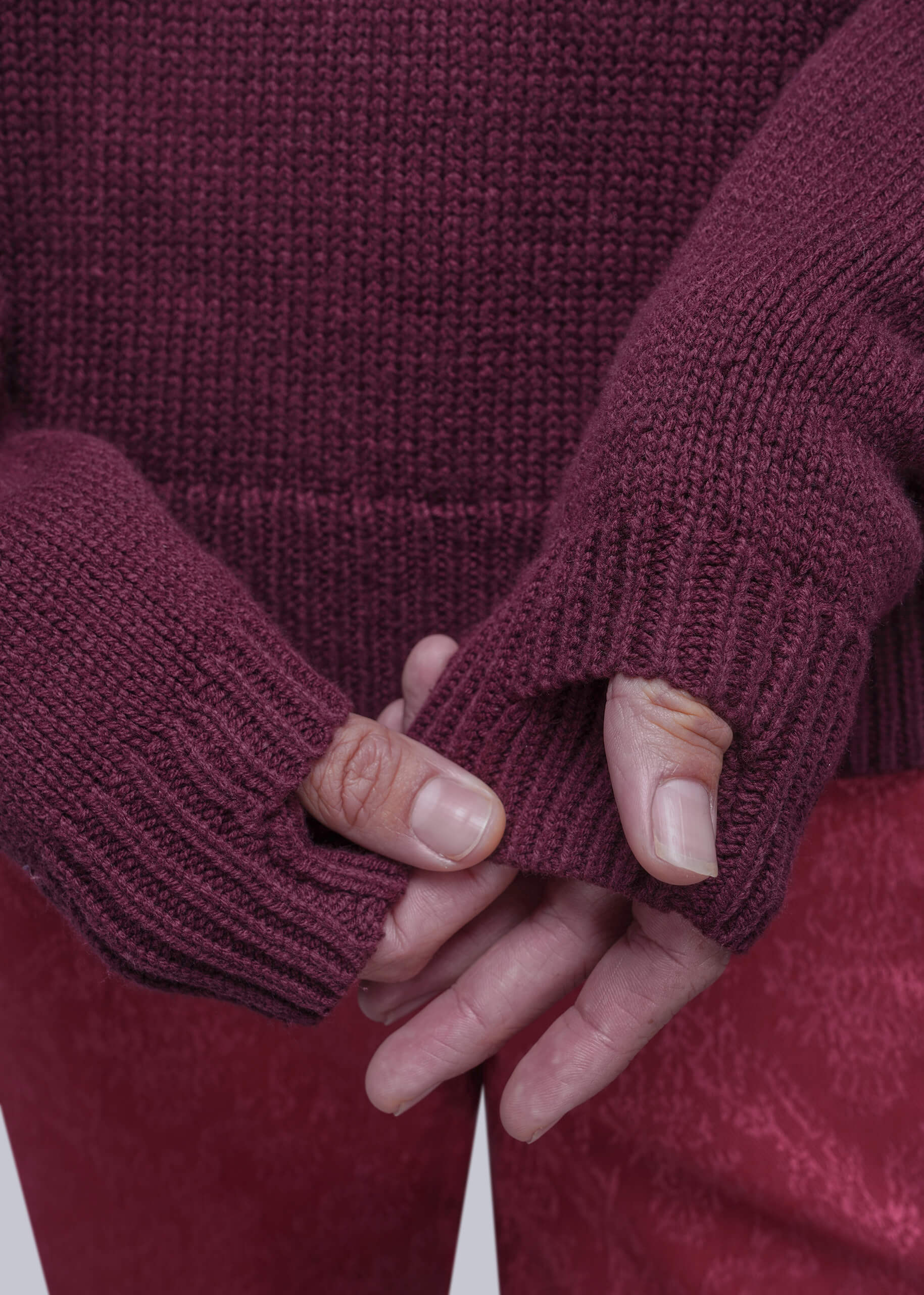 Close-up of a person wearing a chunky knit burgundy sweater with thumbholes and matching burgundy pants, holding their hands together in front of the sweater.
