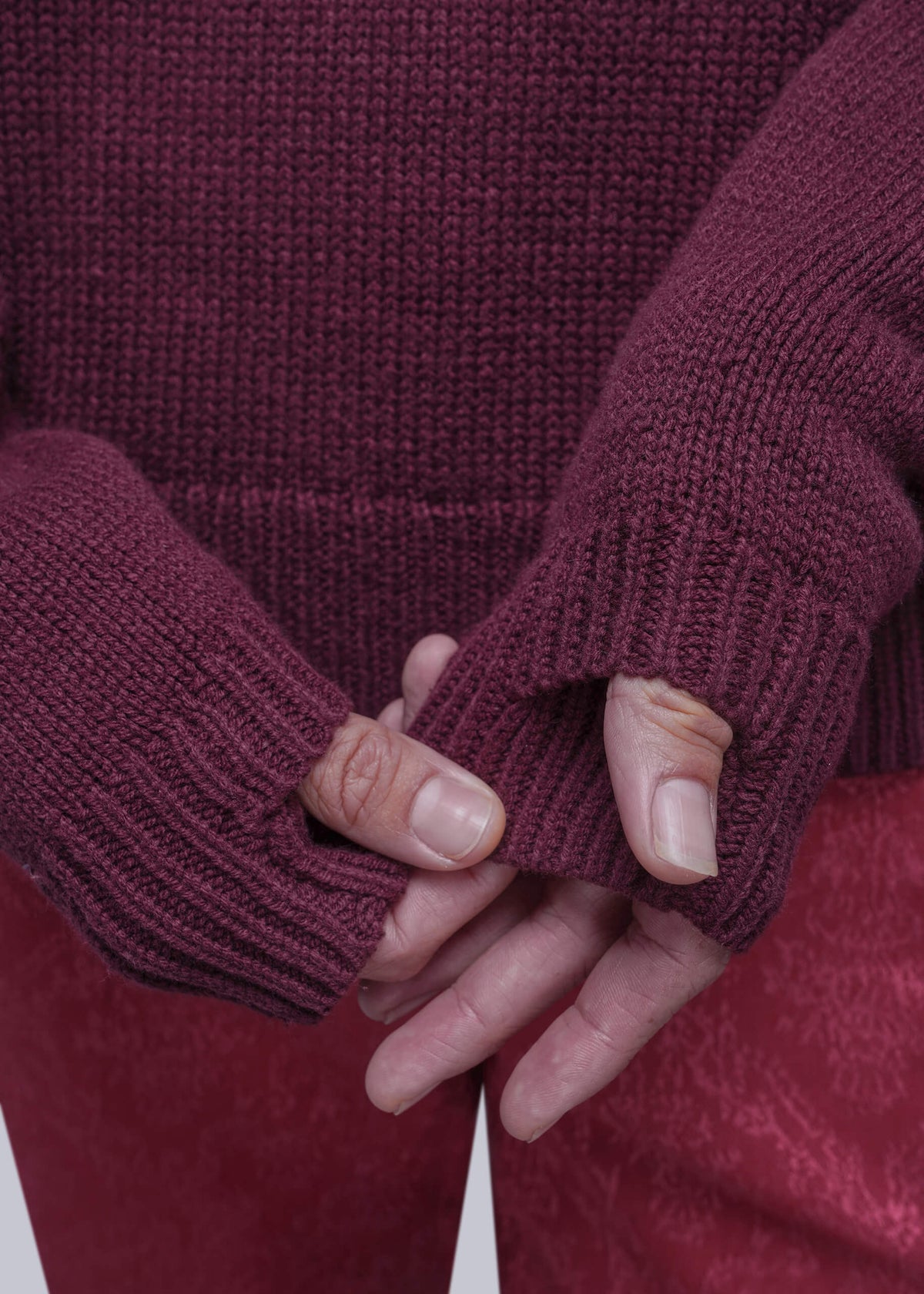 S=Close-up of a person wearing a chunky knit burgundy sweater with thumbholes and matching burgundy pants, holding their hands together in front of the sweater.