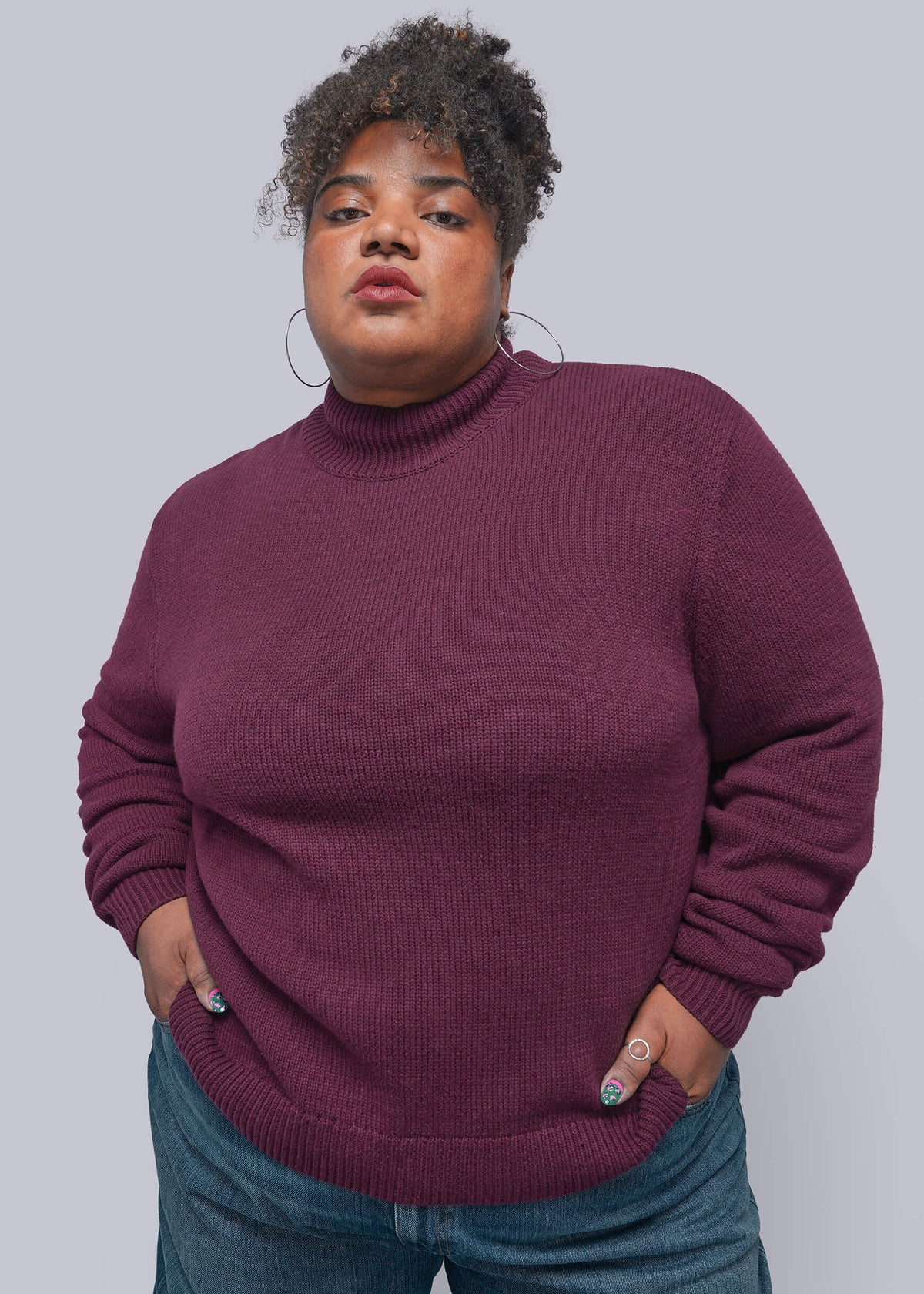1X=A plus-size model wearing a ribbed, long-sleeve mock neck sweater in a deep plum purple color, paired with blue jeans, against a solid gray studio background.